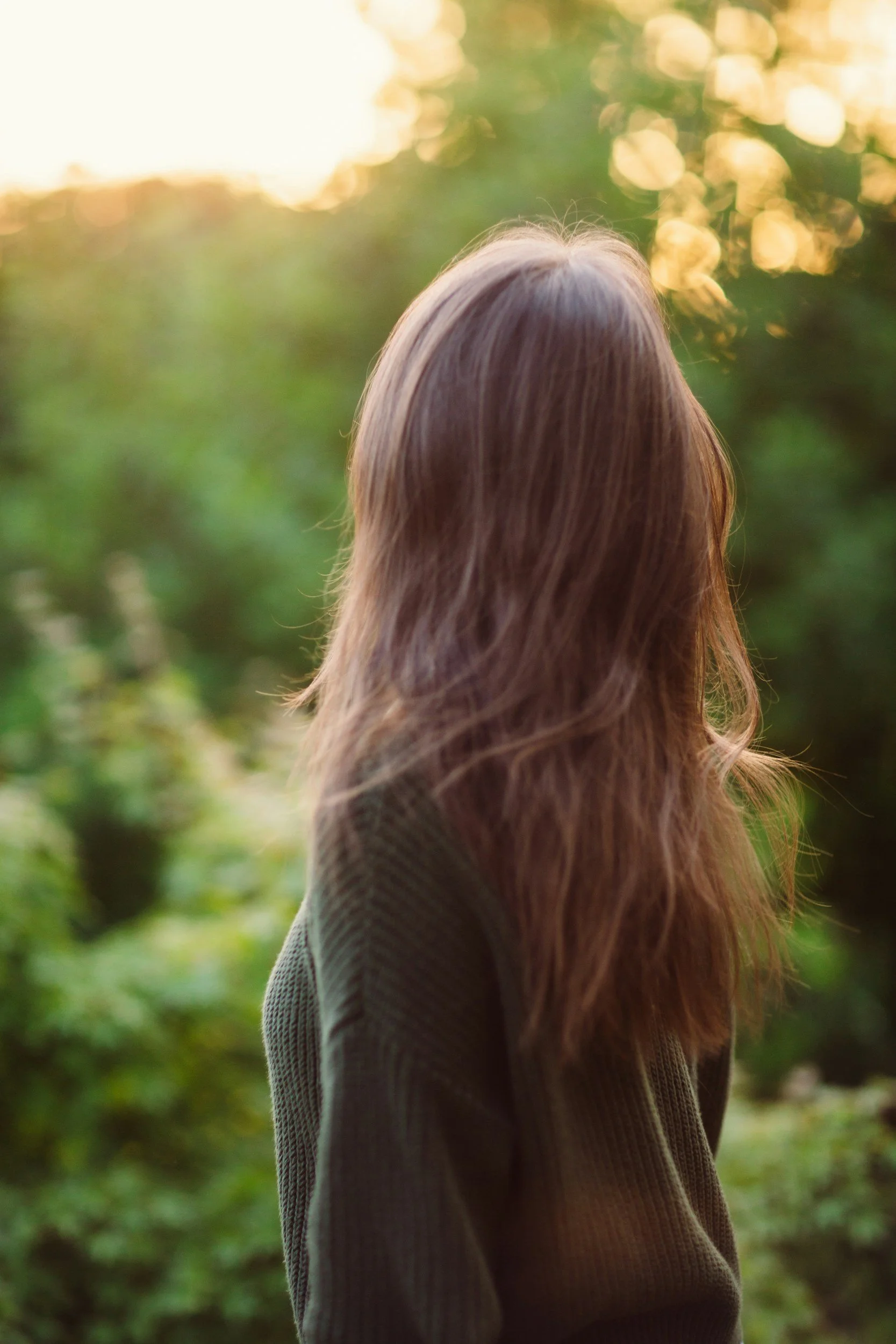A woman with long hair standing outdoors during sunset, facing away from the camera, surrounded by green trees and foliage.