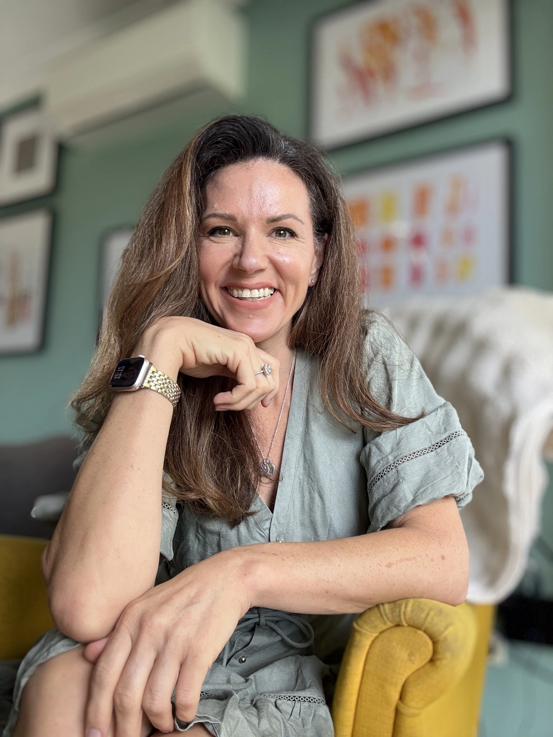 A woman with long brown hair smiling and sitting on a yellow chair in a room with green walls and framed artwork.