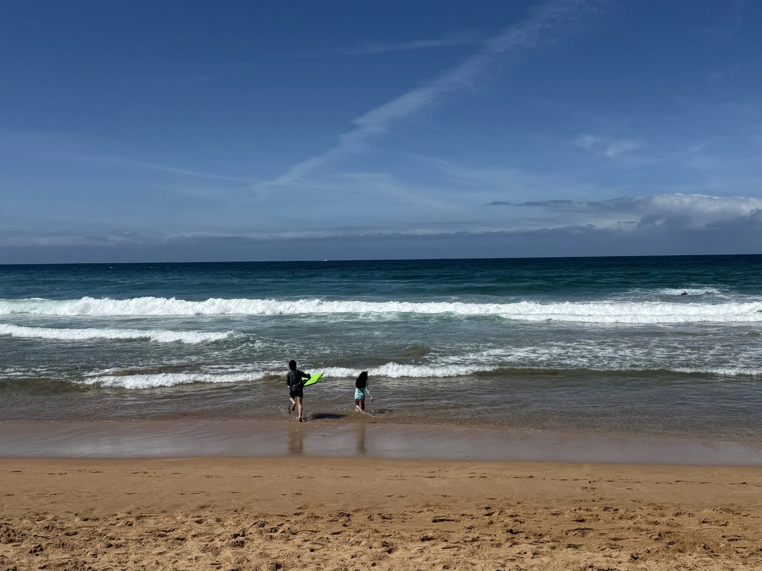 Two children walking into the ocean at the beach, one holding a green surfboard, with sand in the foreground and blue sky with clouds overhead.