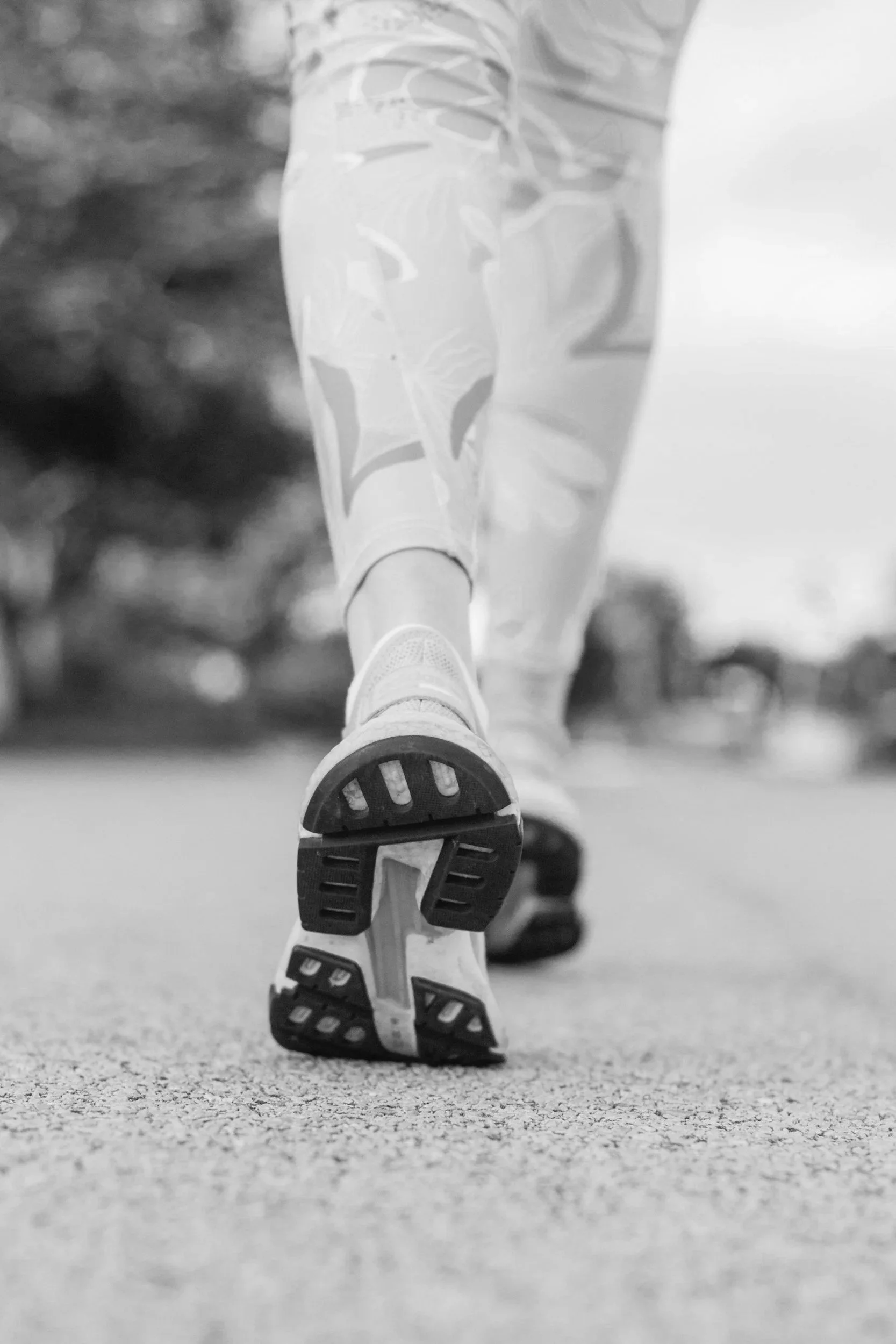 Close-up of a person's legs and feet as they walk or run outdoors, wearing athletic leggings and shoes, on a textured surface with trees in the background.