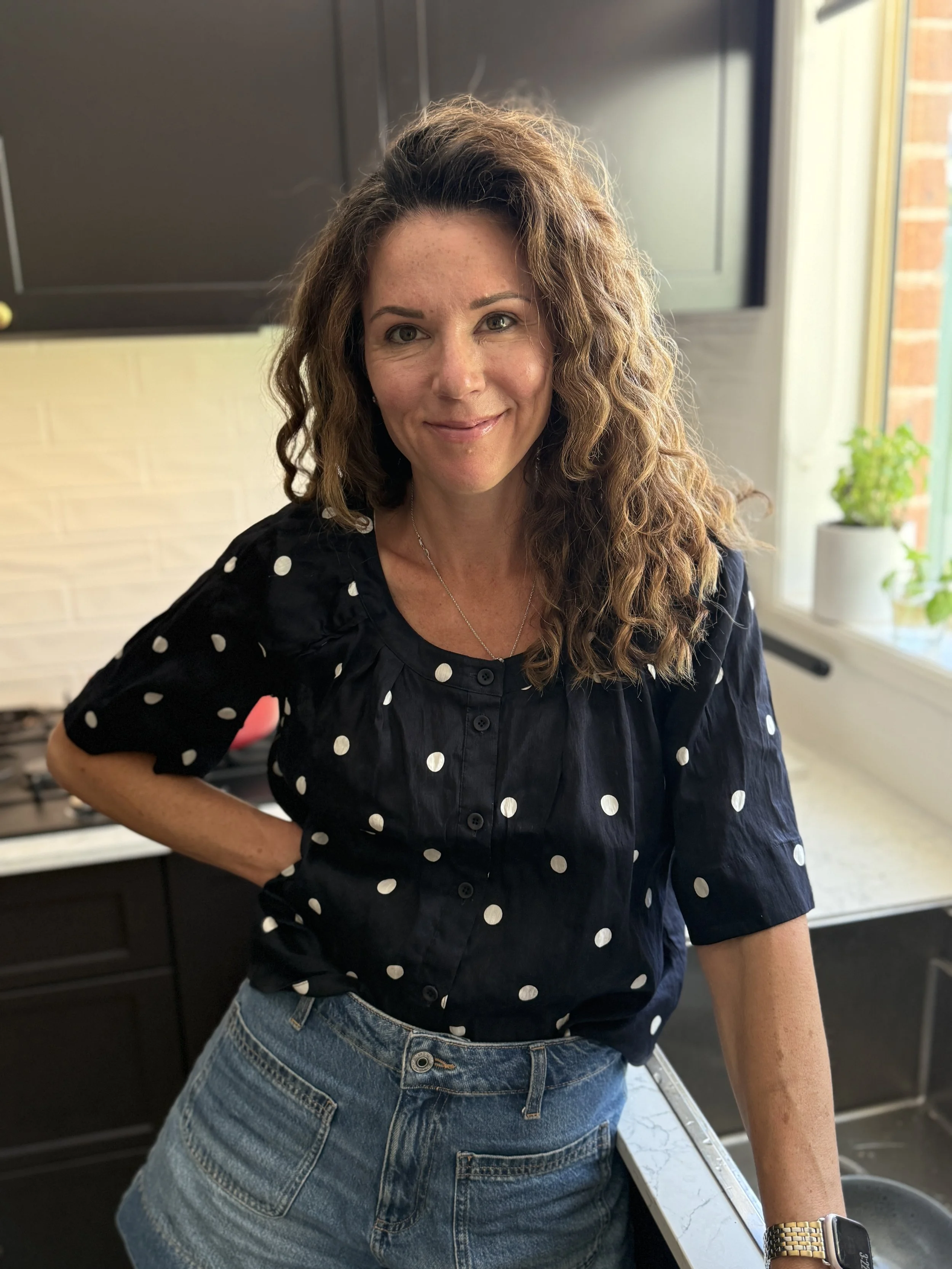 A woman with curly brown hair, wearing a black blouse with white polka dots and blue jeans, standing in a kitchen near a window with a potted plant on the windowsill.