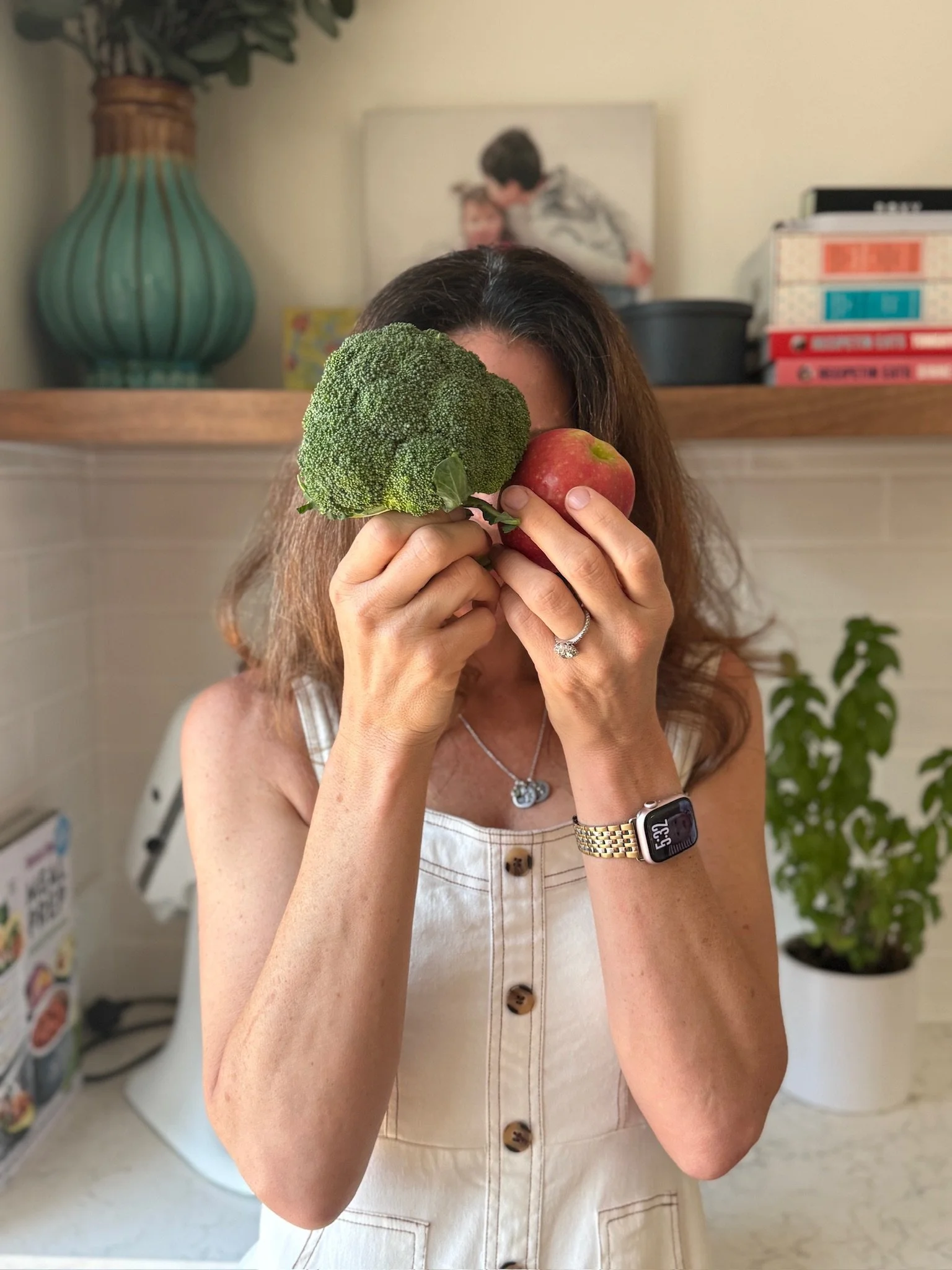 A woman holding a piece of broccoli and an apple in front of her face, with a bouquet of broccoli and apple covering her face, in a kitchen setting.