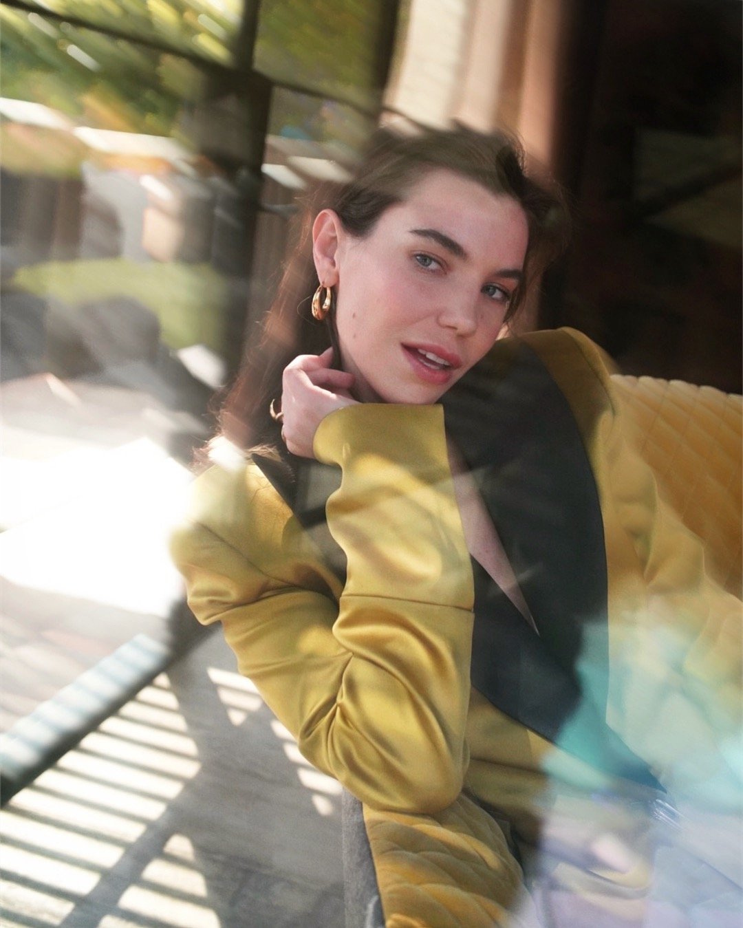 A young woman with brown hair and blue eyes taking a selfie while sitting outside, wearing a yellow jacket and gold earrings, with sunlight and reflections visible.