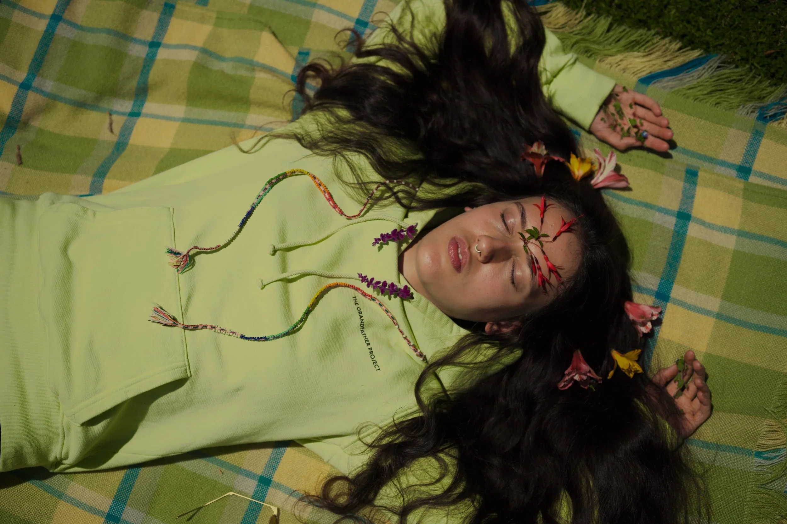 A woman with long dark hair lies on a green plaid blanket with her eyes closed. She is wearing a light green hoodie and has flowers intertwined in her hair and on her face. Two colorful beaded necklaces rest on her chest. The scene is natural and serene.