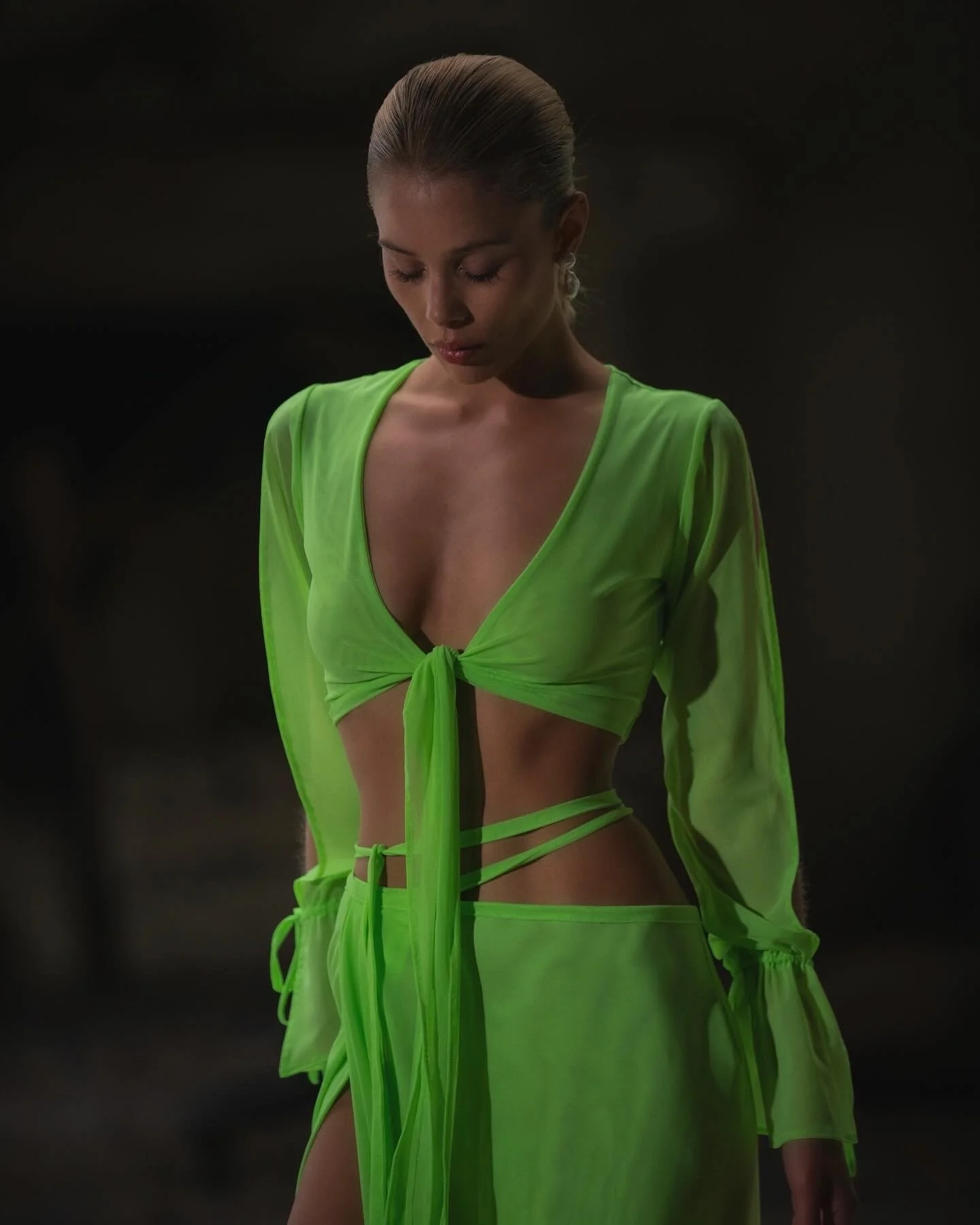 Woman wearing a neon green, sheer two-piece outfit with a crop top and skirts, tied in the front, standing with her eyes closed and head tilted downward, dark background.