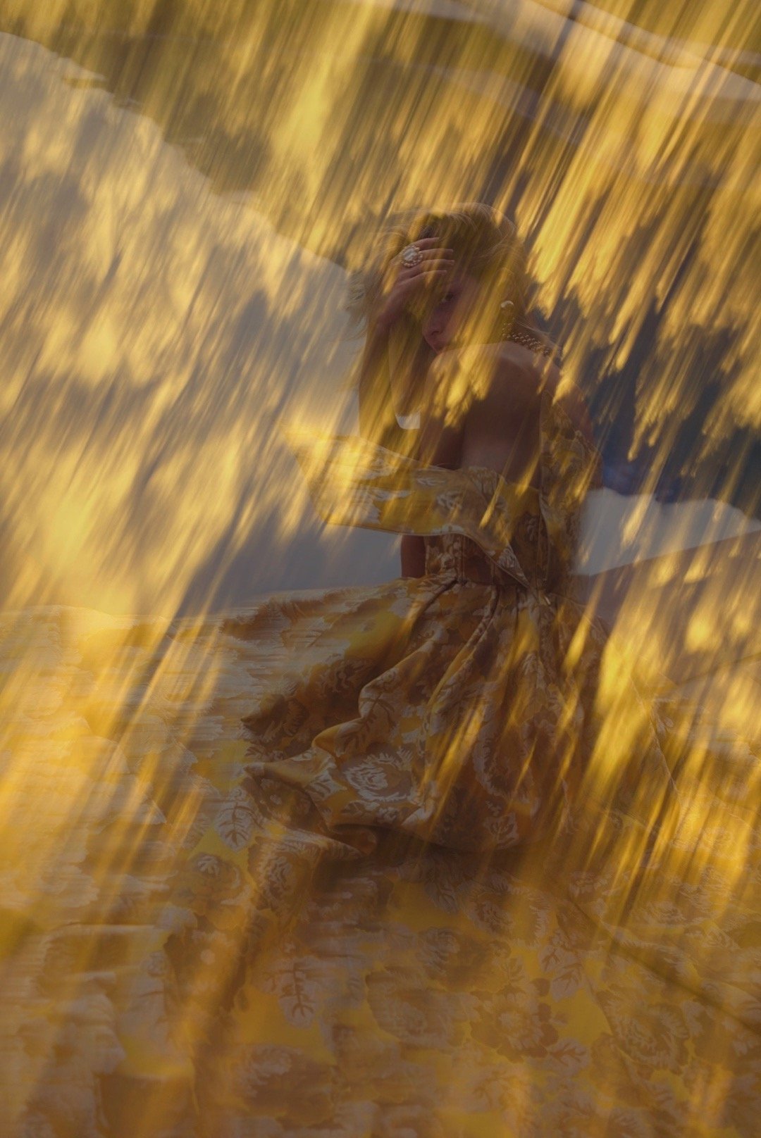 A woman sitting in a yellow floral dress, with her hand on her forehead, reflected in a window with yellow foliage outside.