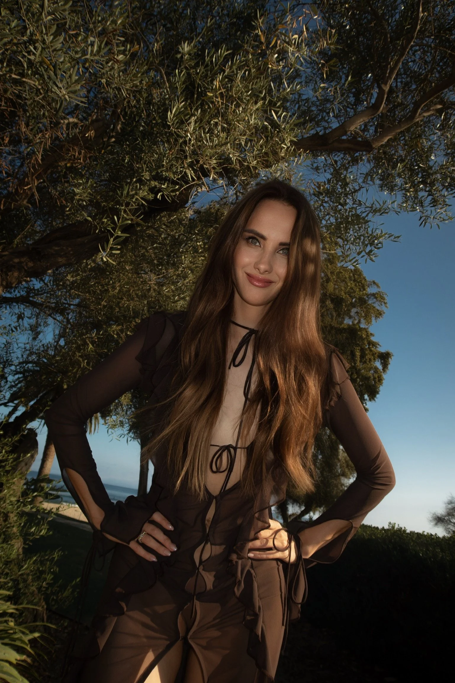 A woman standing outdoors in front of a large tree with lush foliage during twilight, smiling and looking at the camera.