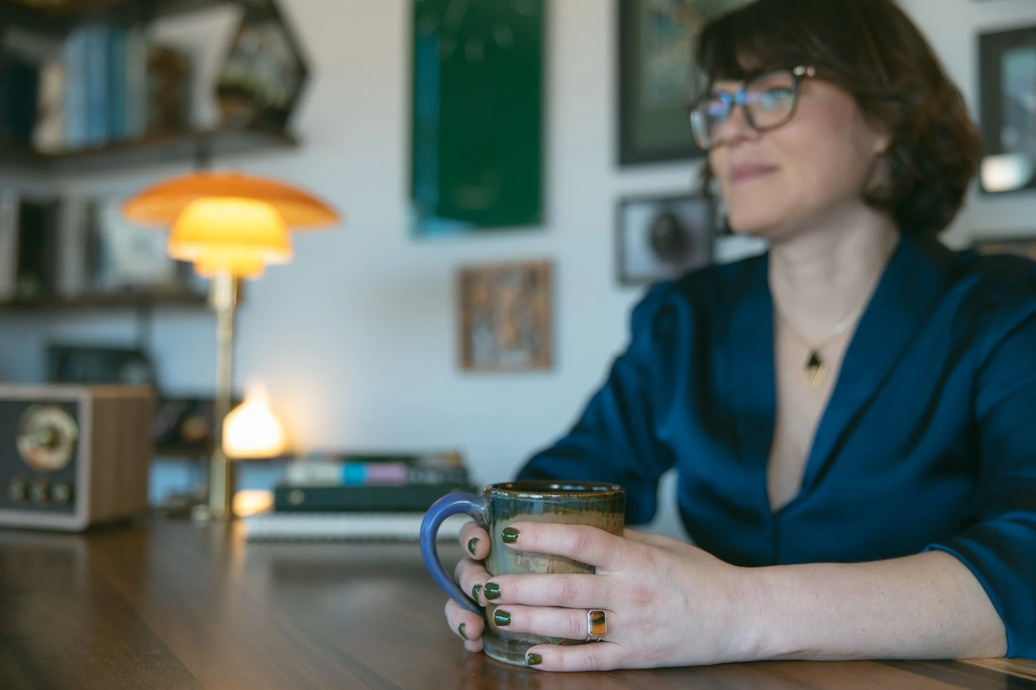 A woman with curly hair and glasses sitting at a desk, holding a mug in her hands, with a blurred background of shelves, books, and a lamp.