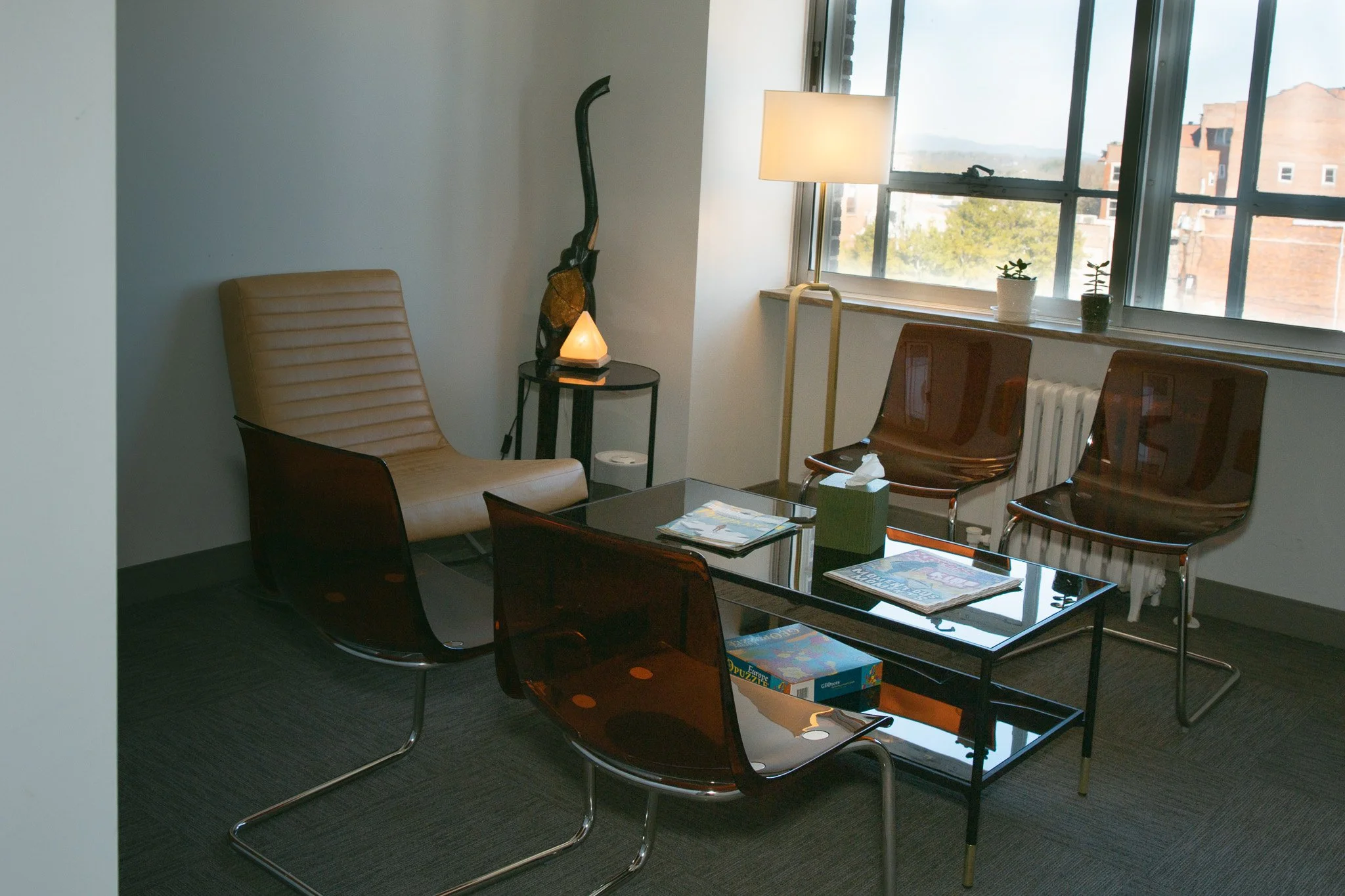 Office room with a tan leather chair, two brown chairs, a black glass coffee table with magazines and a tissue box, a window with two small potted succulents, a white lamp, and a lamp with a salt lamp. Outside the window are red brick buildings and trees.