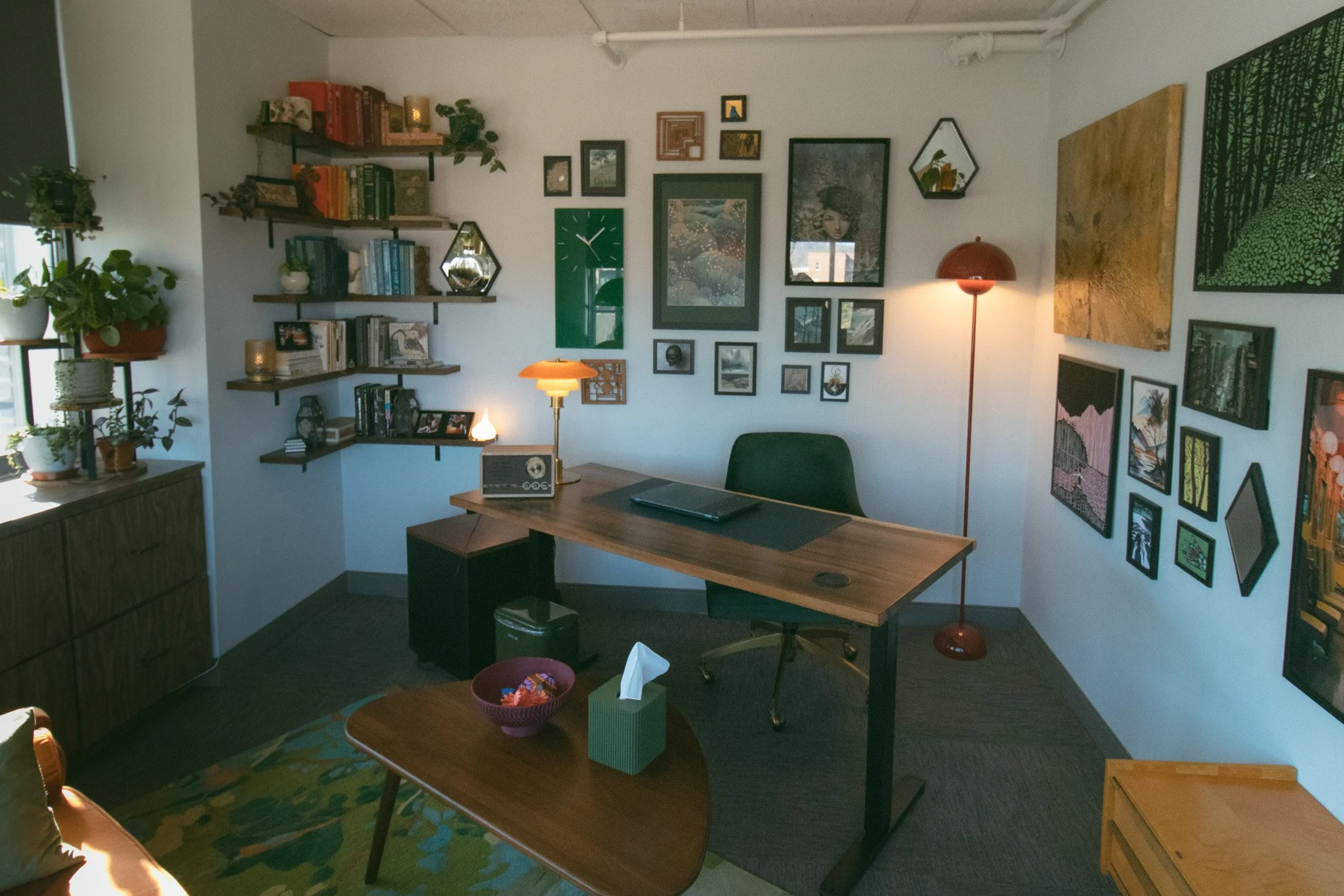 A home office with a wooden desk, green chair, wall art, shelves with books and plants, a lamp, and a window with sunlight.