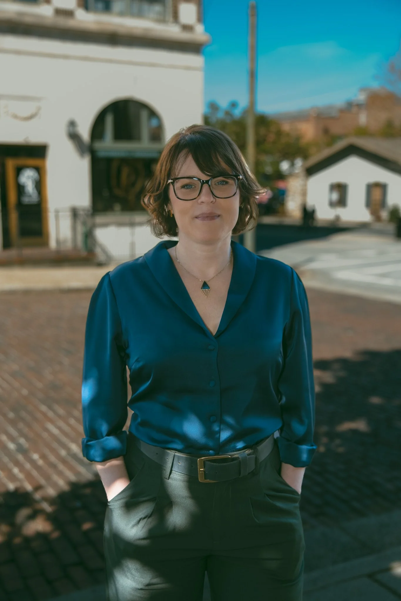 A woman with short brown hair, glasses, and a blue satin blouse standing outdoors on a sunny day with buildings and trees in the background.