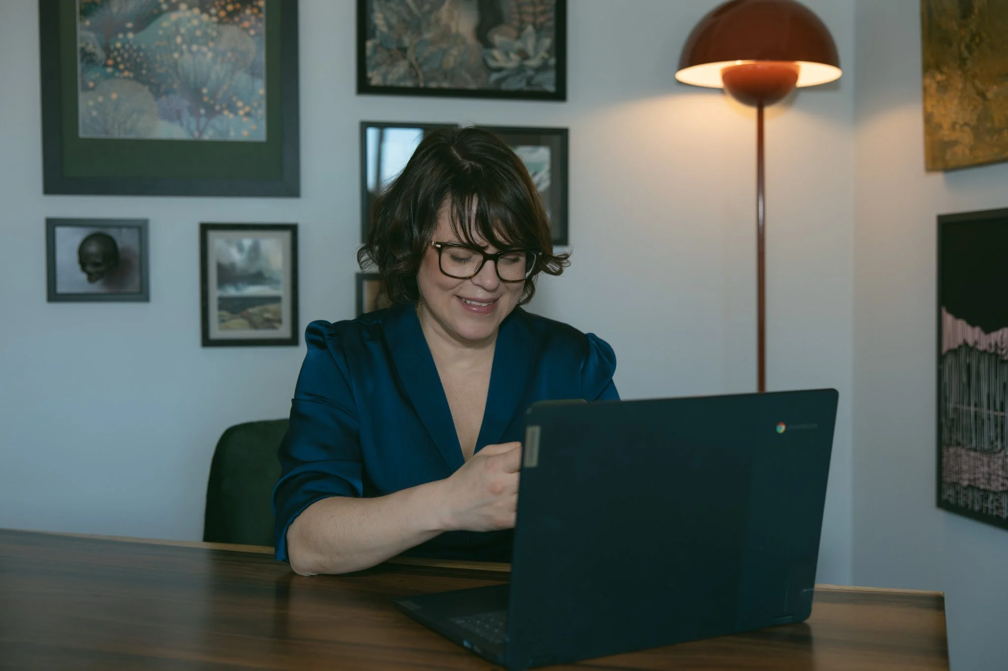 A woman with glasses sitting at a wooden table using a tablet and a laptop, smiling, with framed artwork on the wall behind her and a floor lamp to her right.