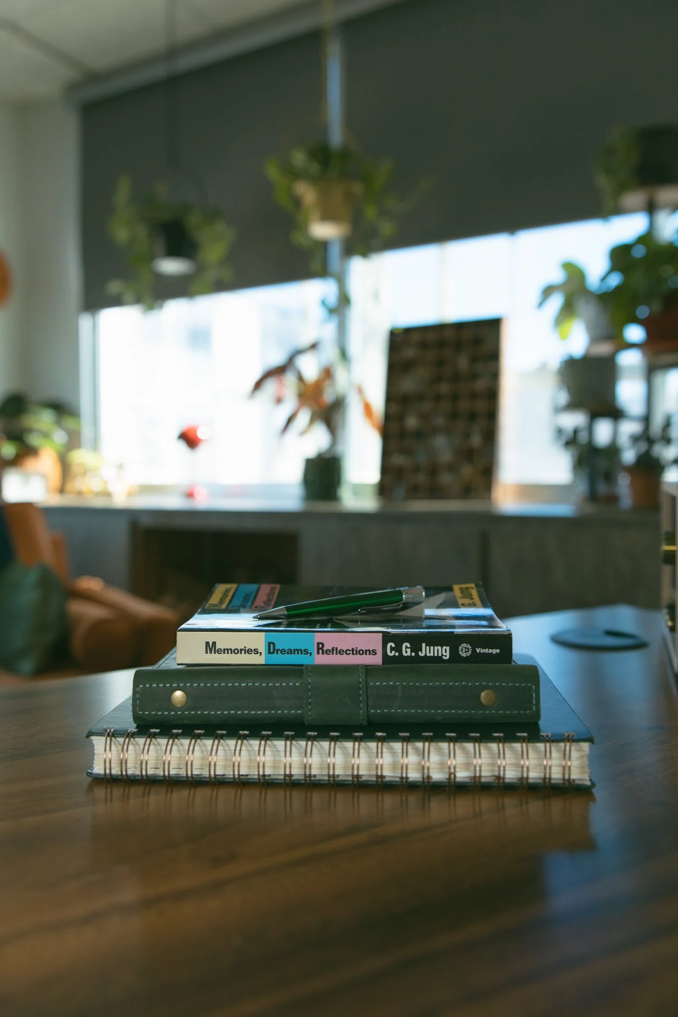 Books, a pen, and a notebook on a desk in a well-lit room with plants and large windows in the background.