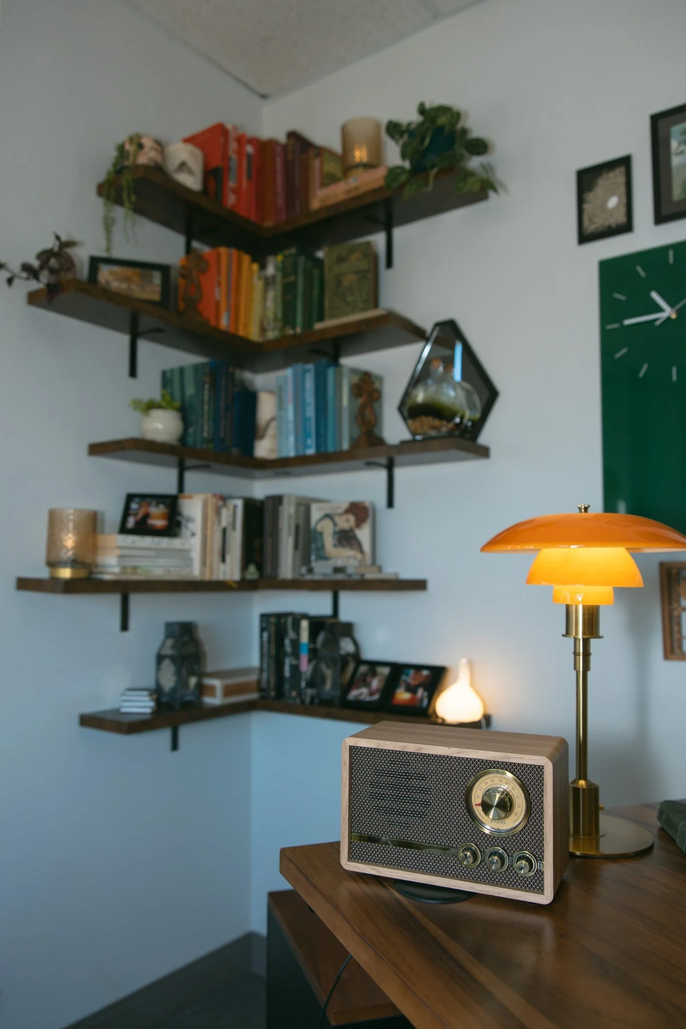 A close-up of a vintage radio and a table lamp on a wooden table in a room with wall-mounted shelves filled with books and decorative items.