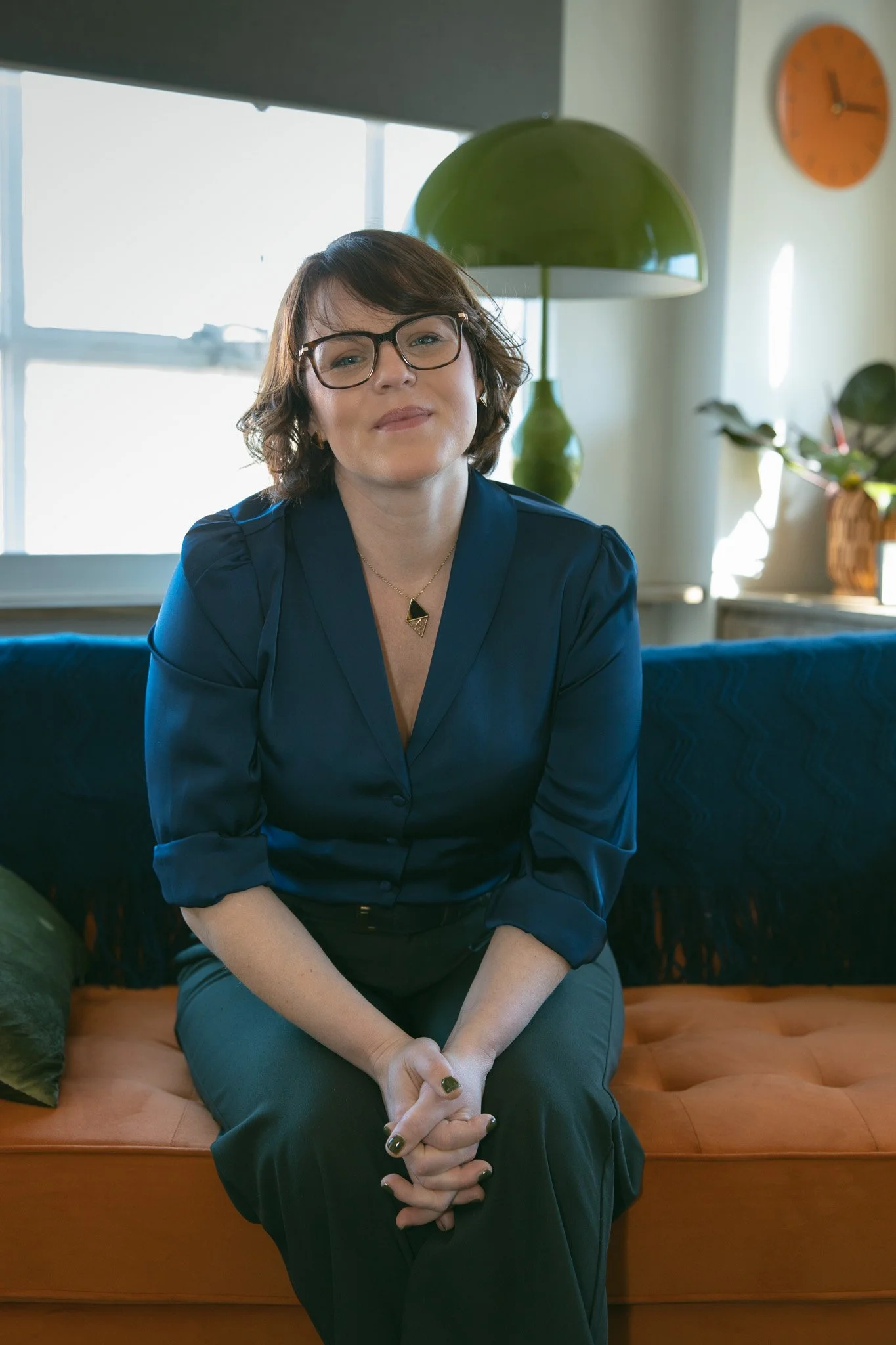 A woman with short brown hair, glasses, and a blue blouse sitting on an orange couch in a well-lit living room with a green lamp, clock, and potted plant in the background.