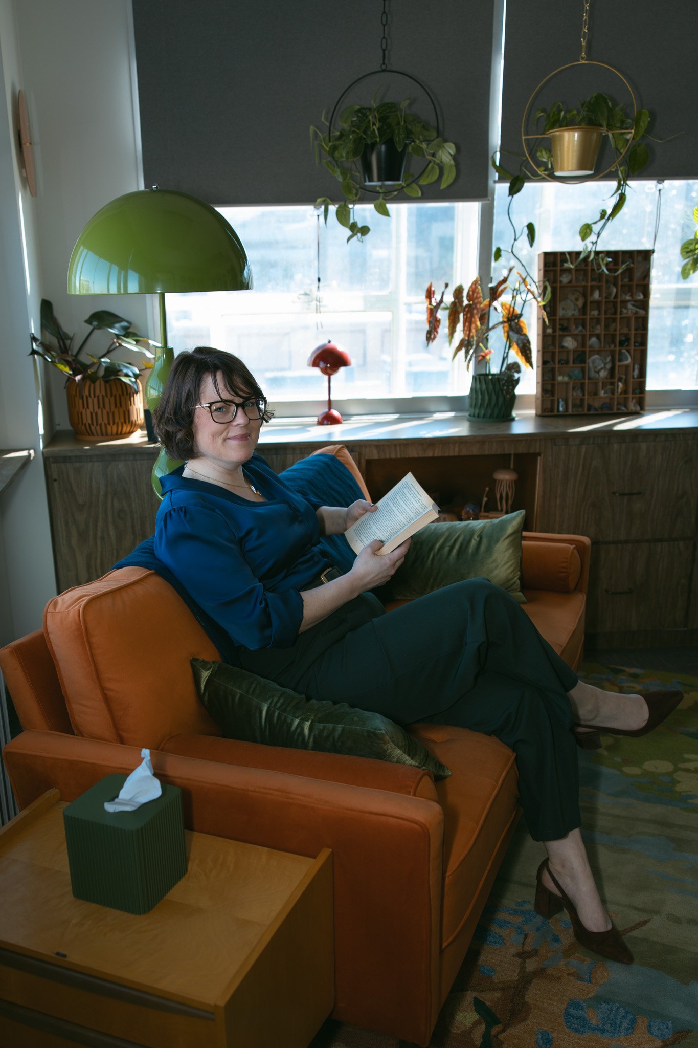 A woman with glasses and chin-length dark hair sits on an orange couch in a well-lit room, reading a book. The room features houseplants, a green desk lamp, and large windows with blinds, with a patterned rug on the floor.