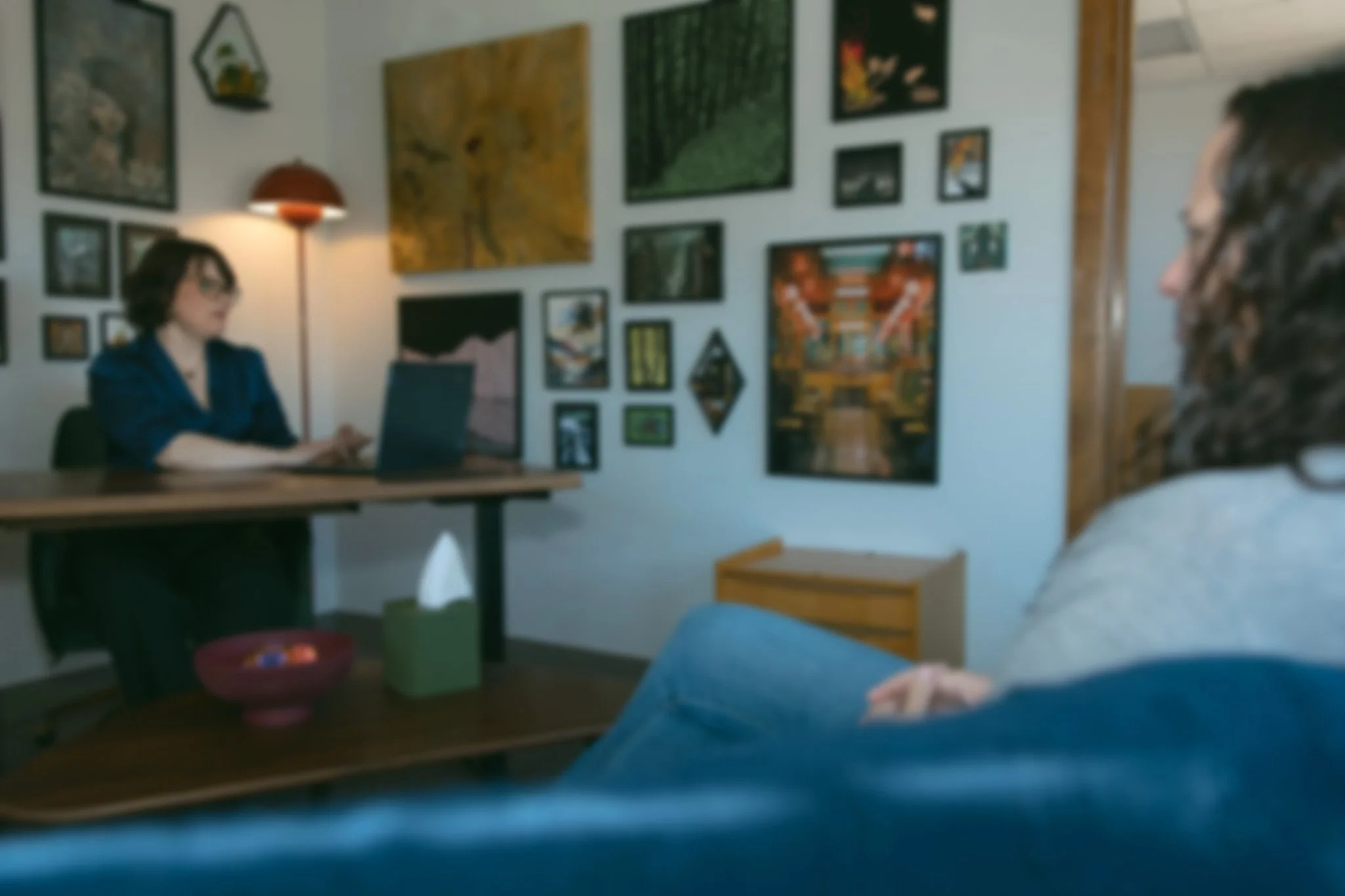 A woman sitting at a desk with a laptop in an art-filled office, as a person with curly hair and glasses sitting on a sofa is seen in the foreground.