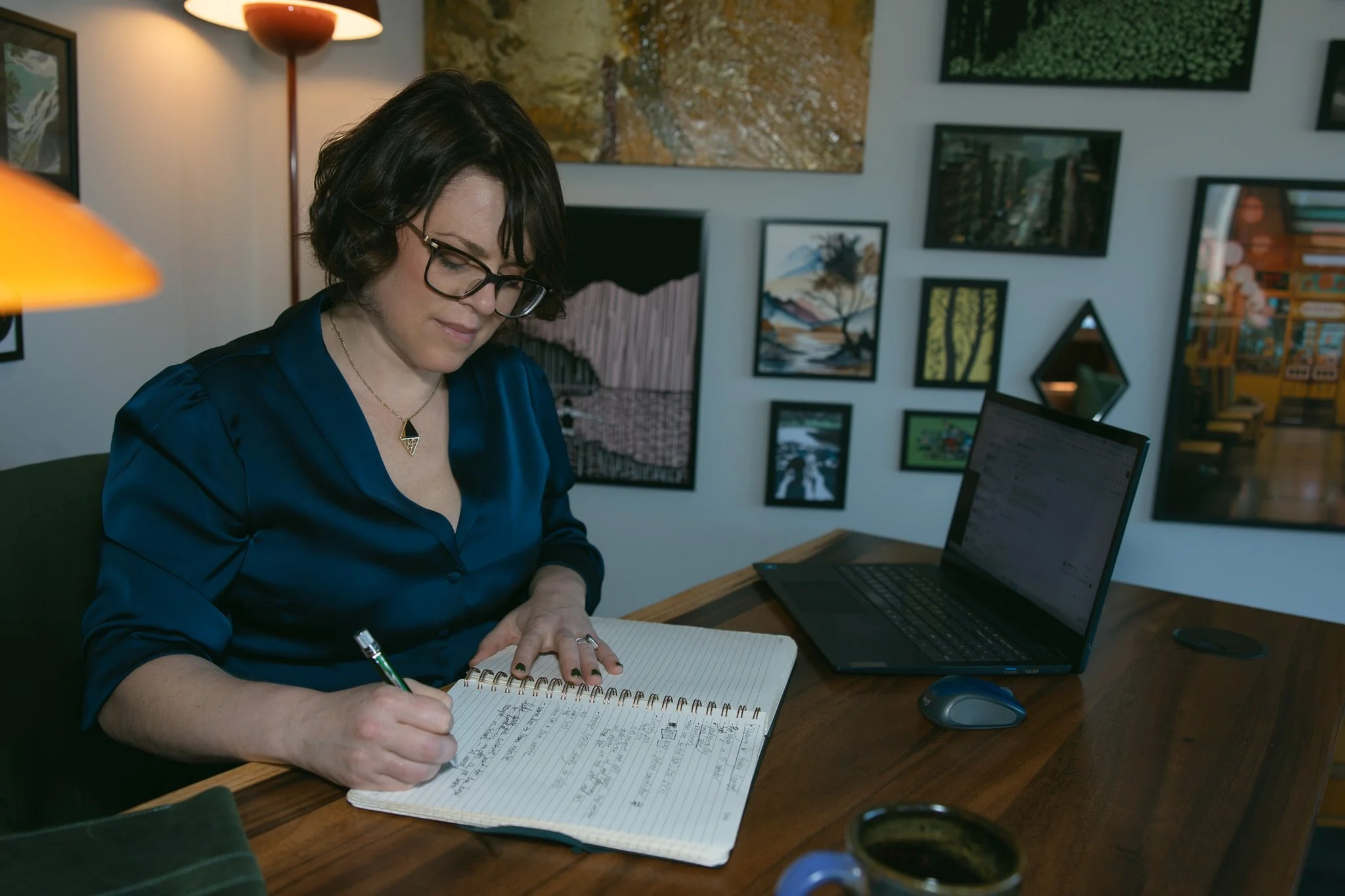 A woman with short dark hair, glasses, and a blue silk blouse sitting at a wooden desk, writing in a notebook. There is a laptop, mouse, and mug on the desk, with framed artwork on the wall behind her.