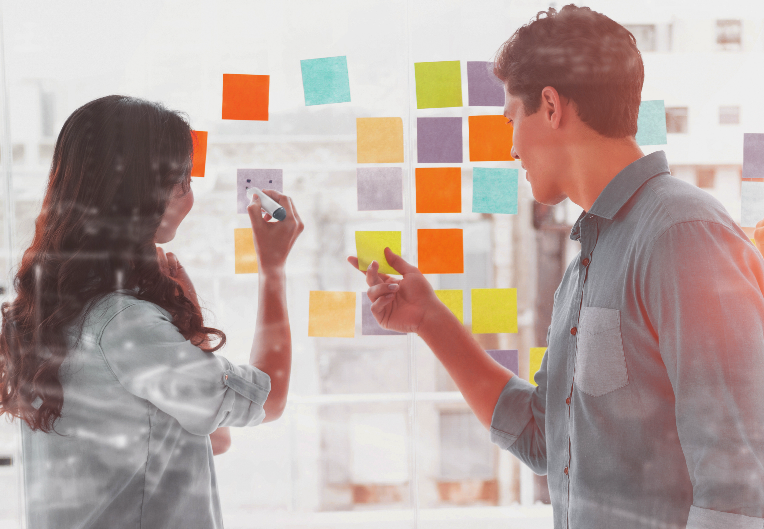 Two people, a woman and a man, are working together on a glass wall covered with colorful sticky notes in an office environment.