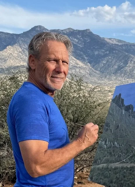 A man in a blue t-shirt painting a Plein Air landscape of mountains and rocky terrain on a canvas outdoors, with mountains and sky in the background.