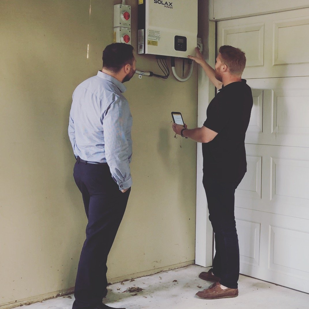 Two men inspecting a solar power inverter mounted on a wall in a garage.