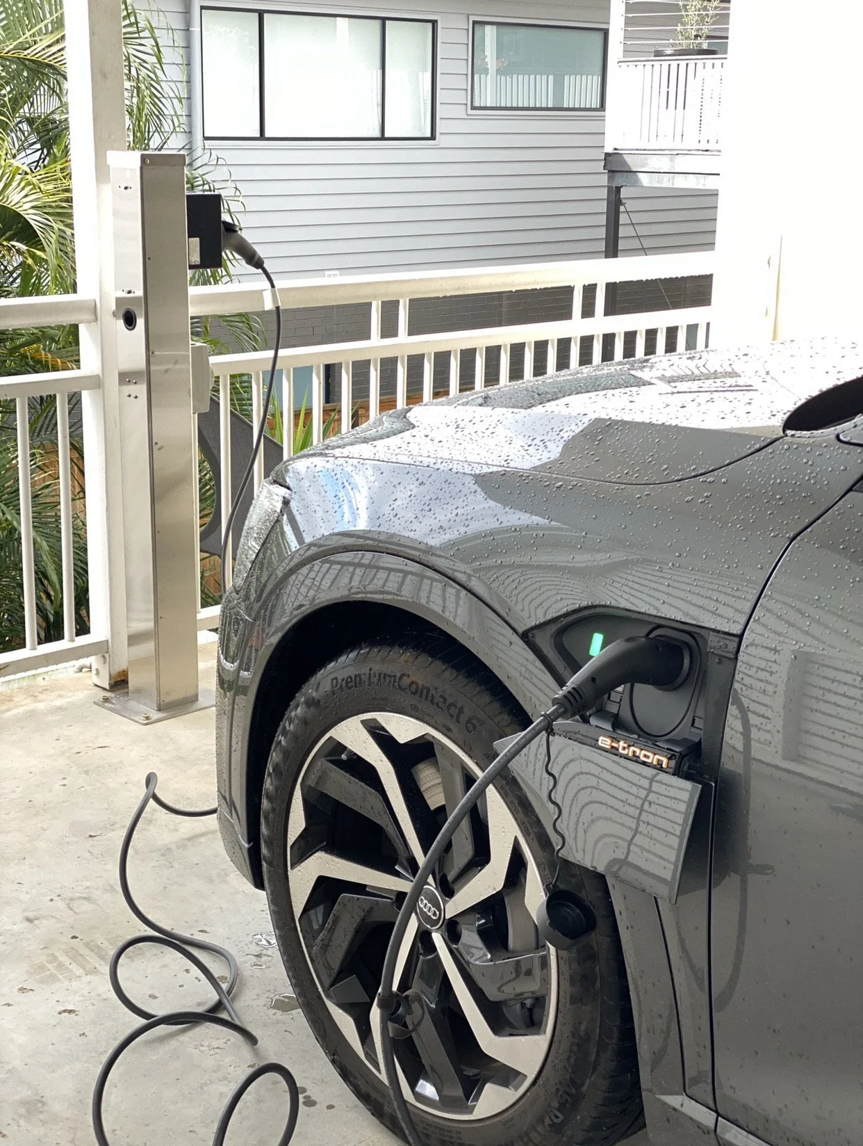 An electric car is charging at a charging station on a parking deck. The car is gray, wet with rain, and connected to two charging cables.