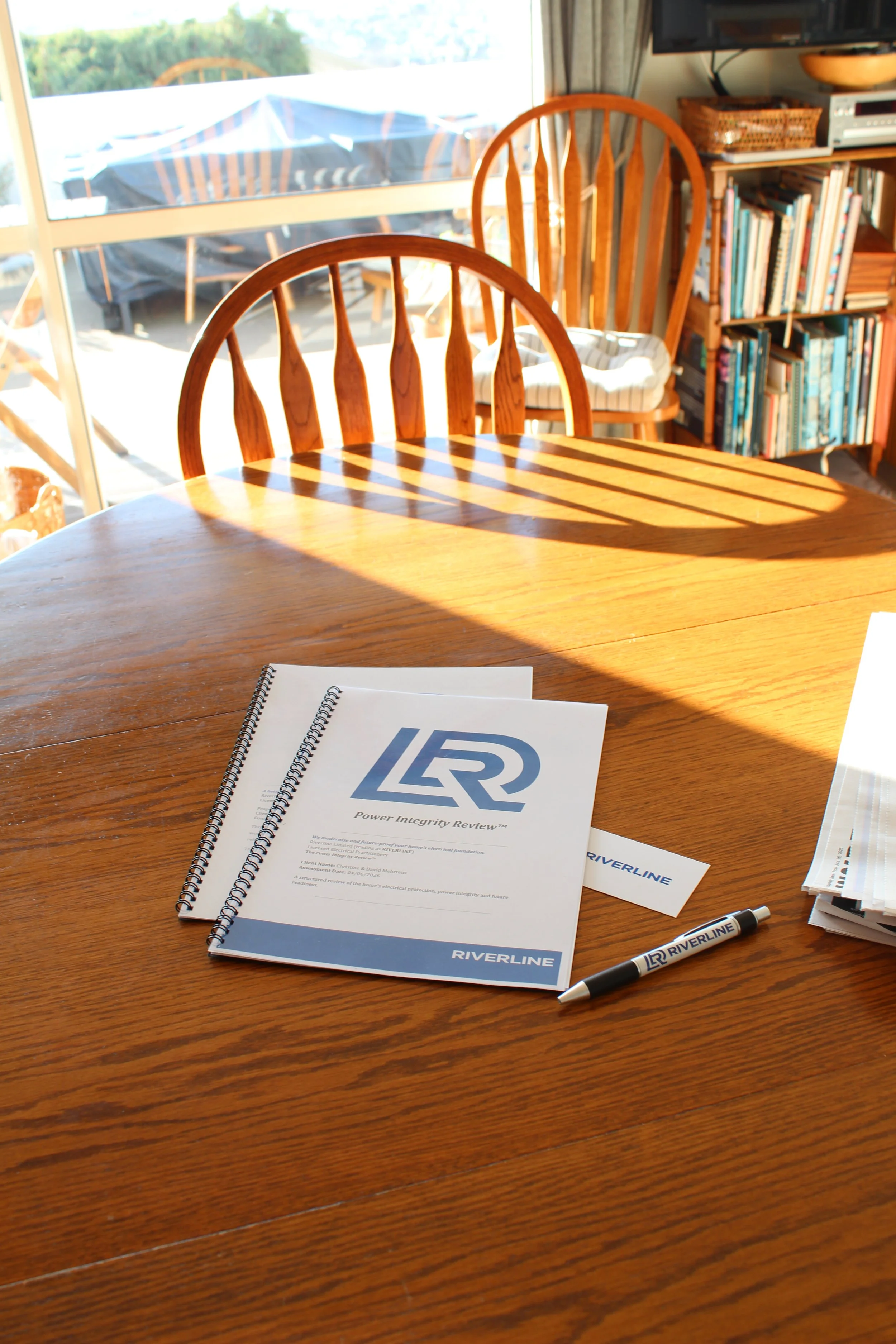 A wooden table with a spiral-bound booklet titled 'Power Integrity Review,' a marked pen, and some additional pamphlets or booklets. In the background, sunlight streams through large windows illuminating the room, which contains wooden chairs and bookshelves filled with books and a TV.