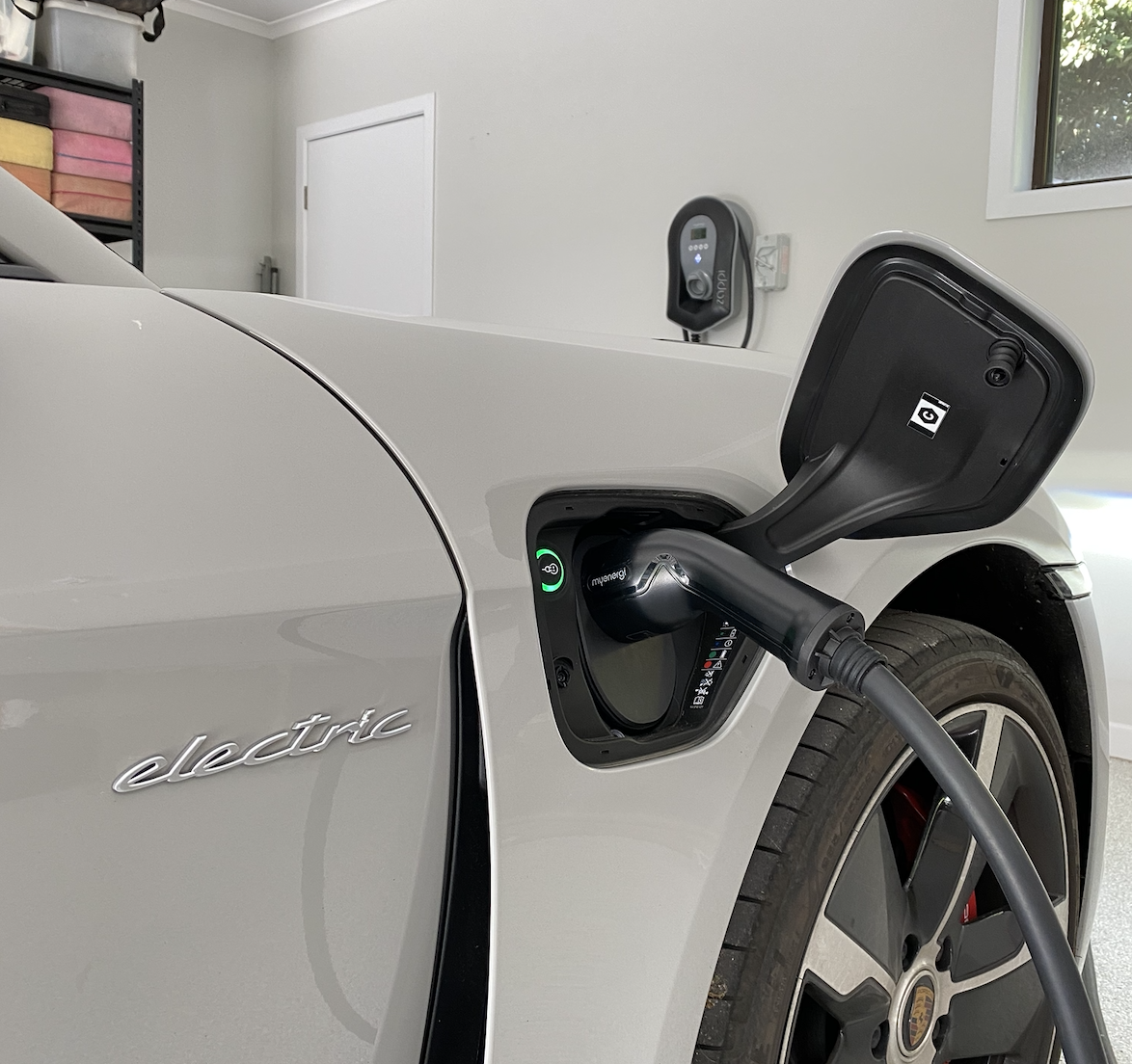Close-up of a white electric Porsche Taycan being charged at a home garage, with a charging cable plugged into the front left port. The car's side badge reads 'electric' and a digital charging monitor is visible.