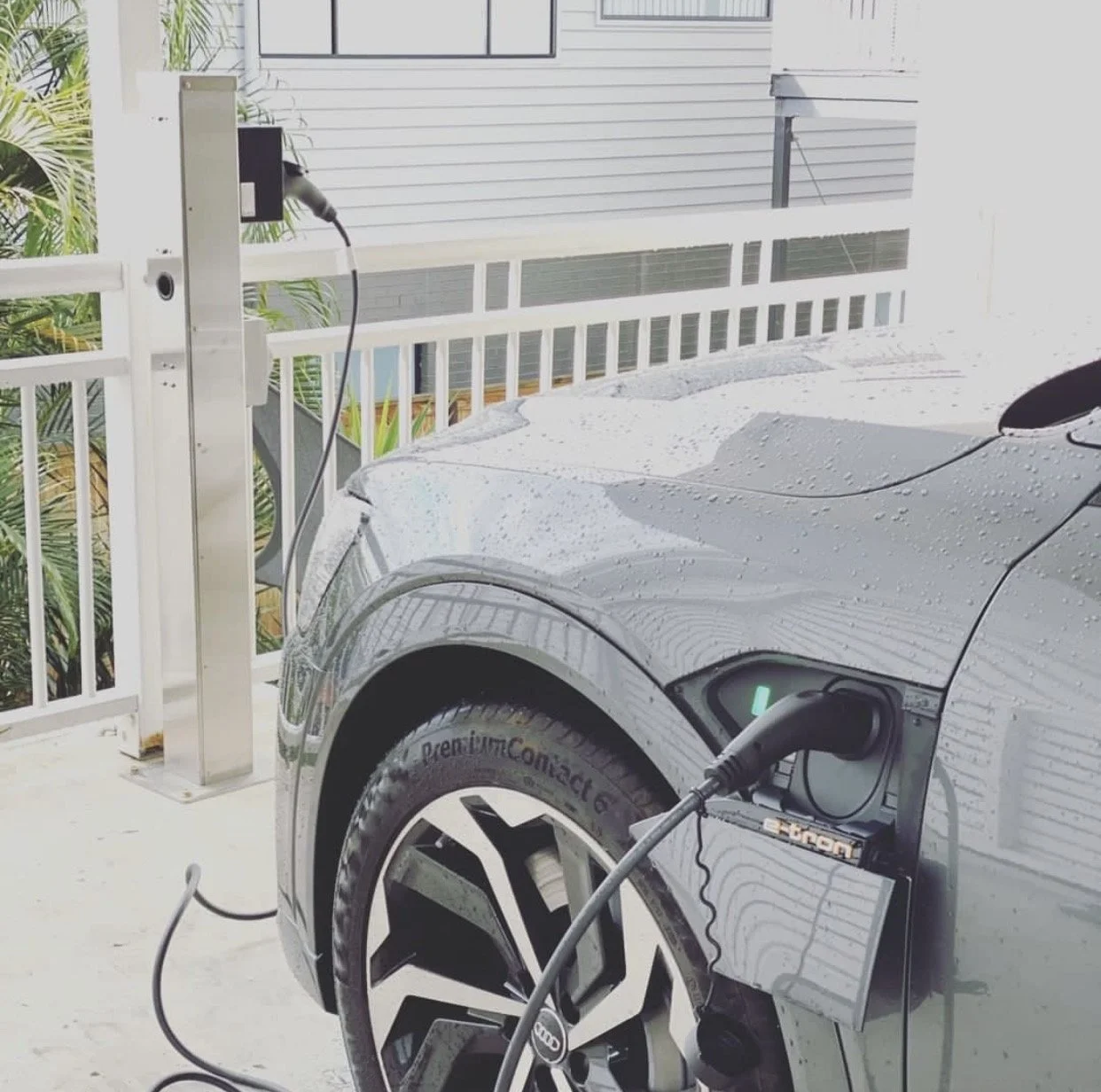 An electric car connected to a charging station on a balcony with rain droplets on its surface and a white railing in the background.