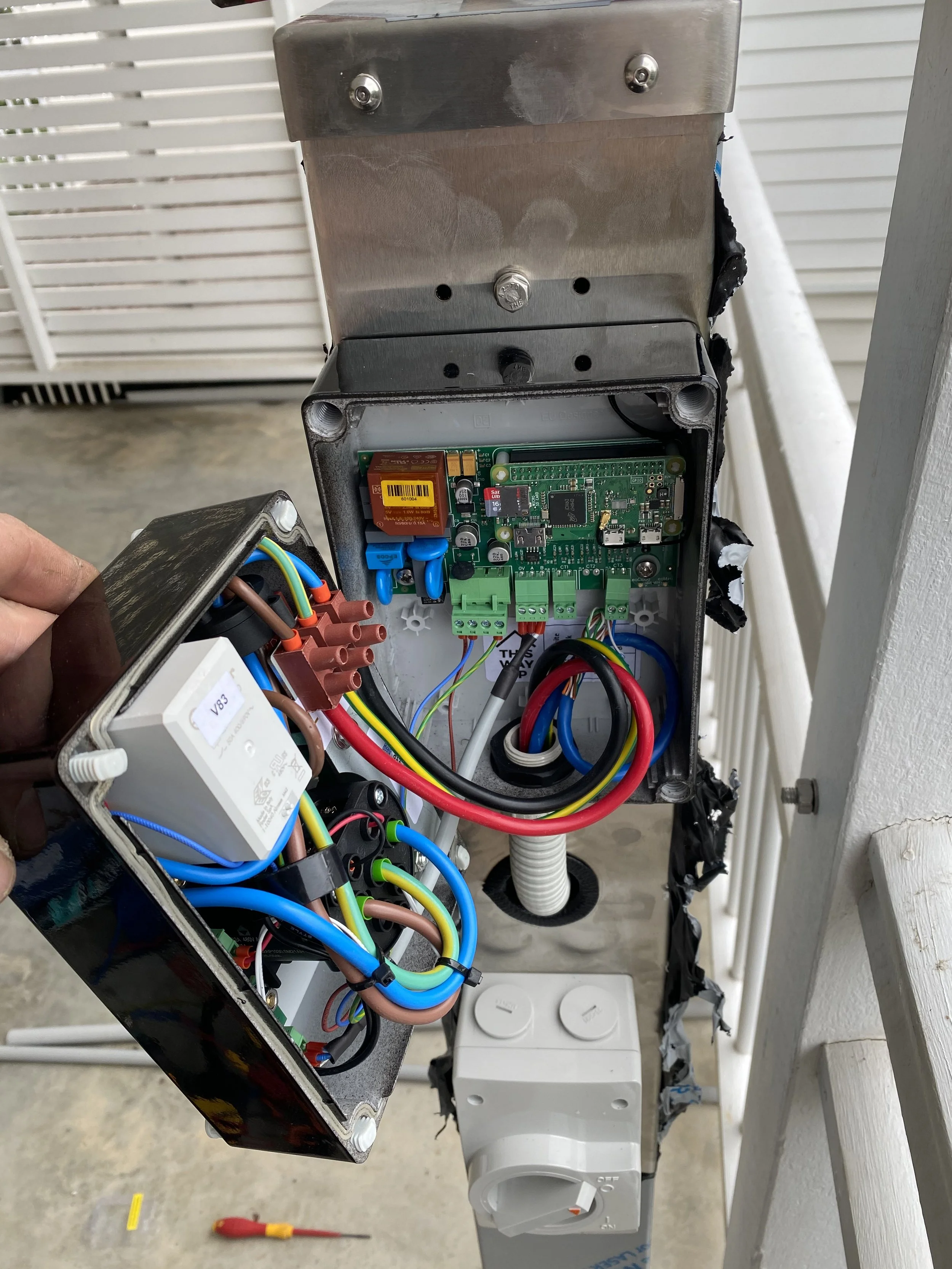 Open electrical box with various wires, circuit boards, and components, with a hand holding a smaller box connected to the system. A power outlet is visible below.