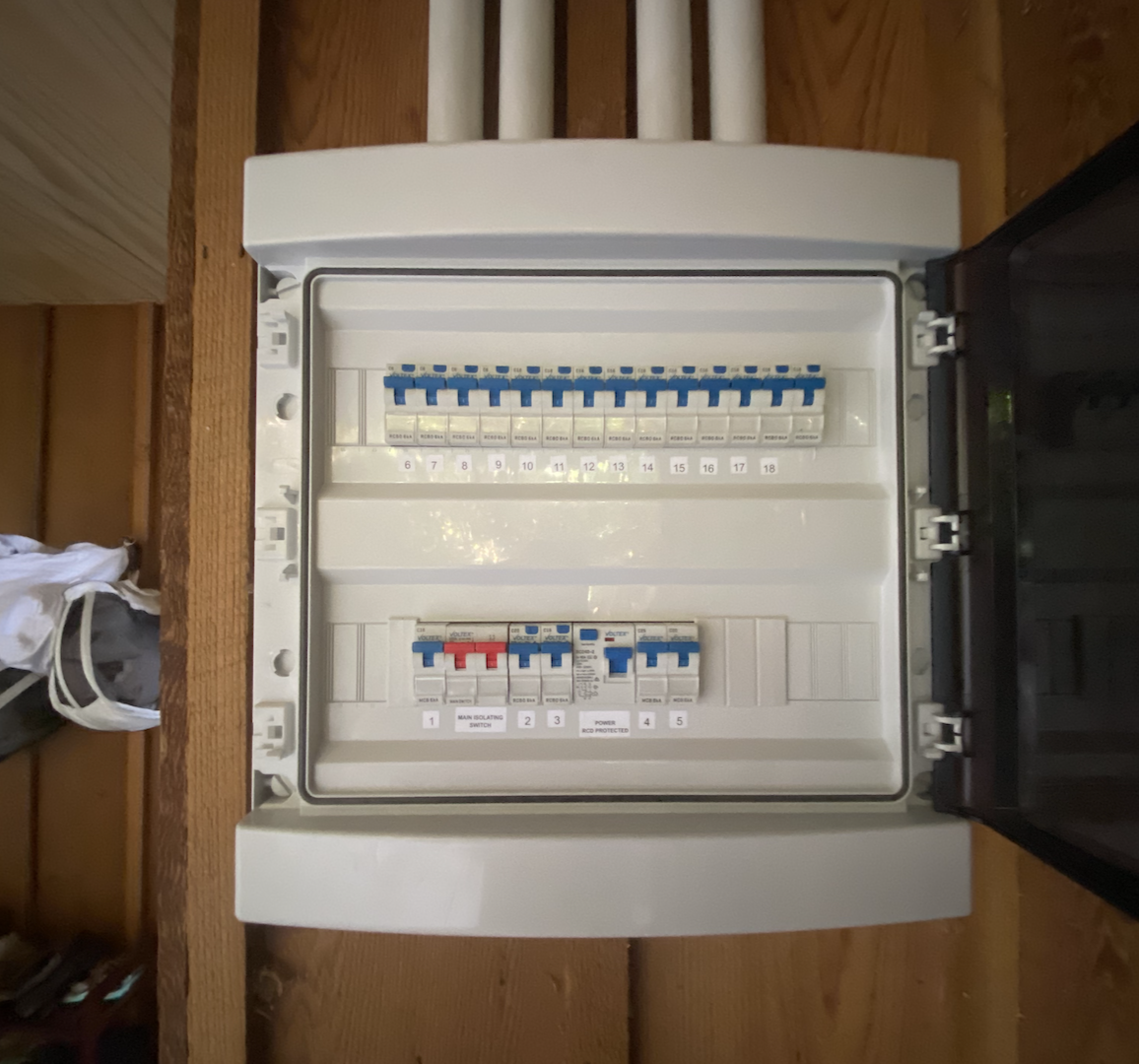 An electrical breaker panel with multiple circuit breakers, mounted on a wooden wall, with some clothing items visible to the left.