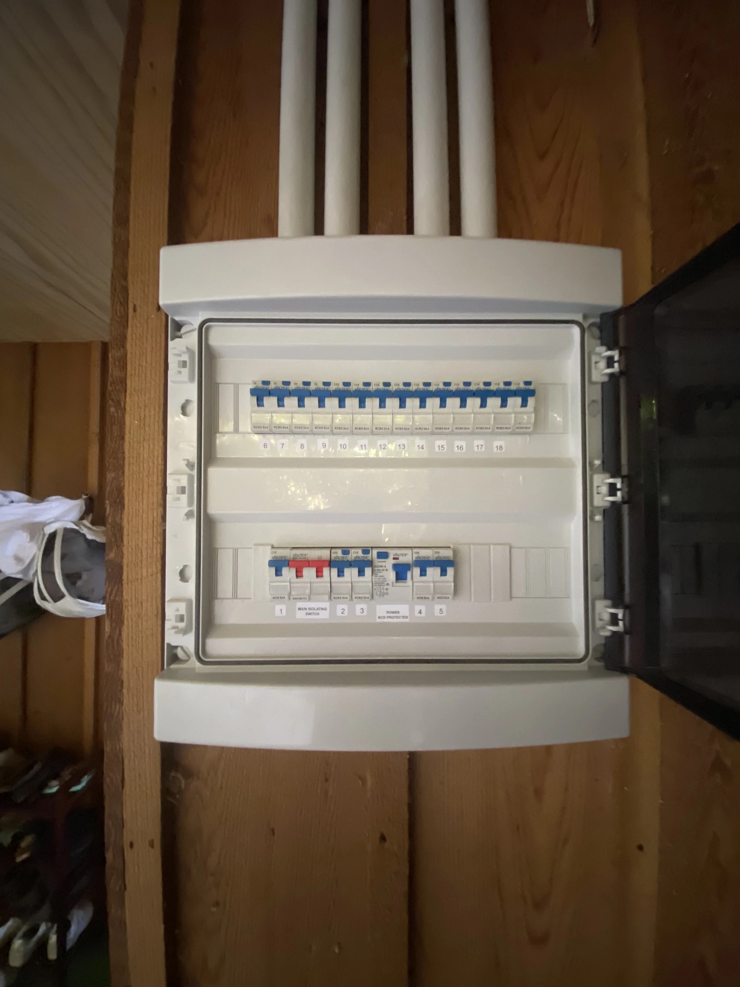 Electrical breaker box mounted on a wooden wall with a transparent cover open, showing circuit breakers inside.