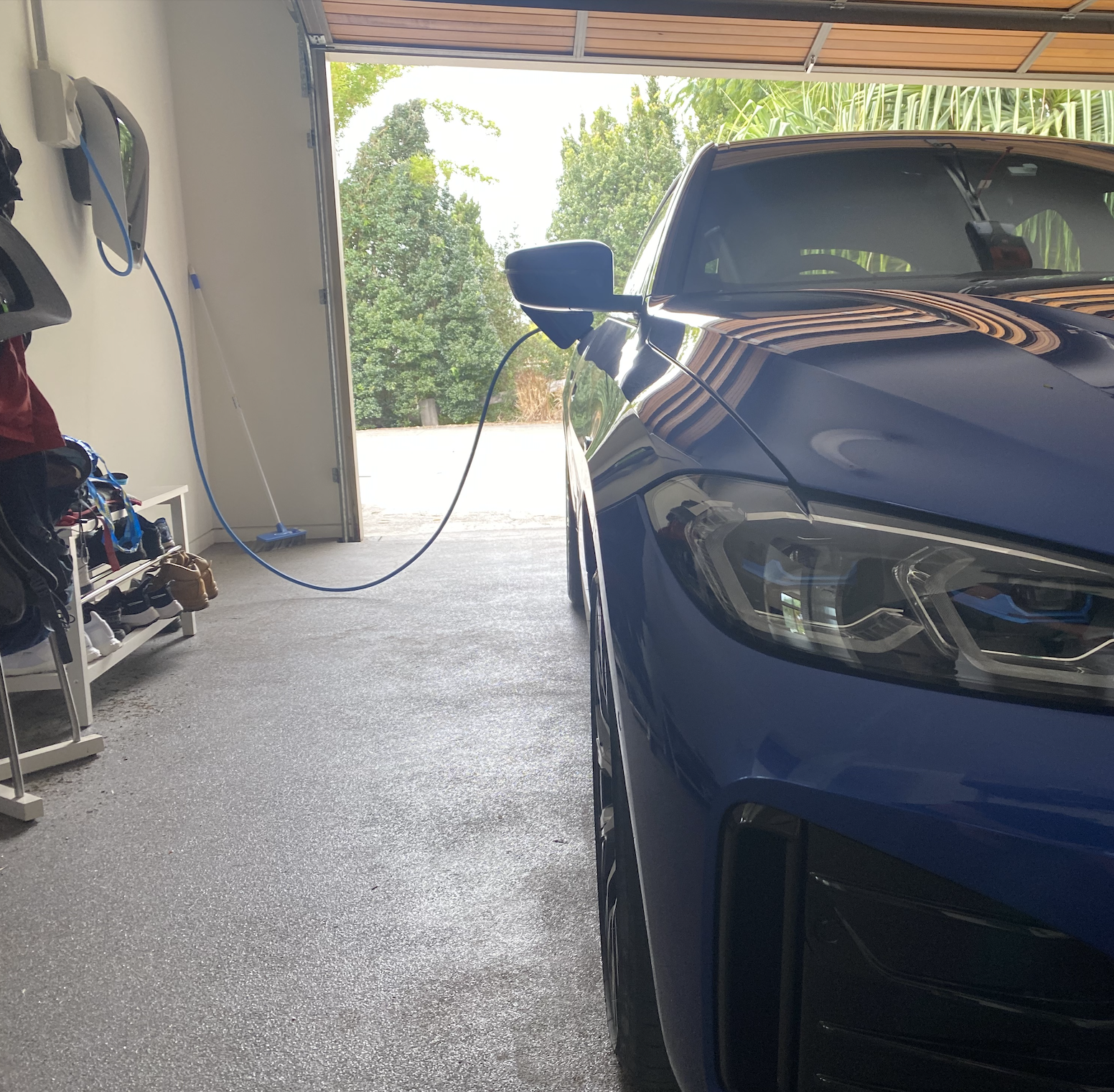 Blue electric car plugged into a charger inside a garage with green trees visible outside.
