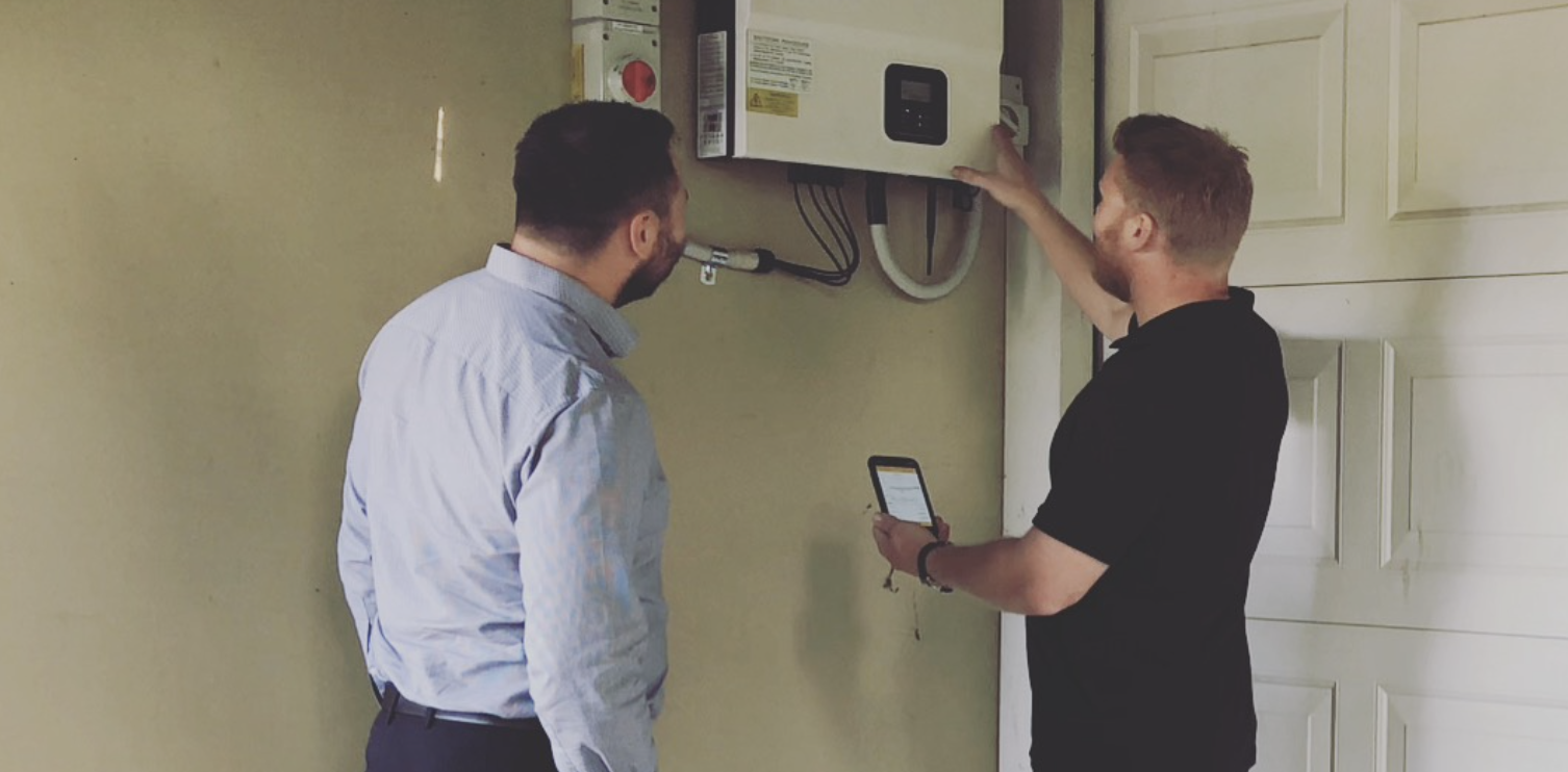 Two men inspecting an electrical control panel, one is holding a smartphone and the other is pointing at the panel.