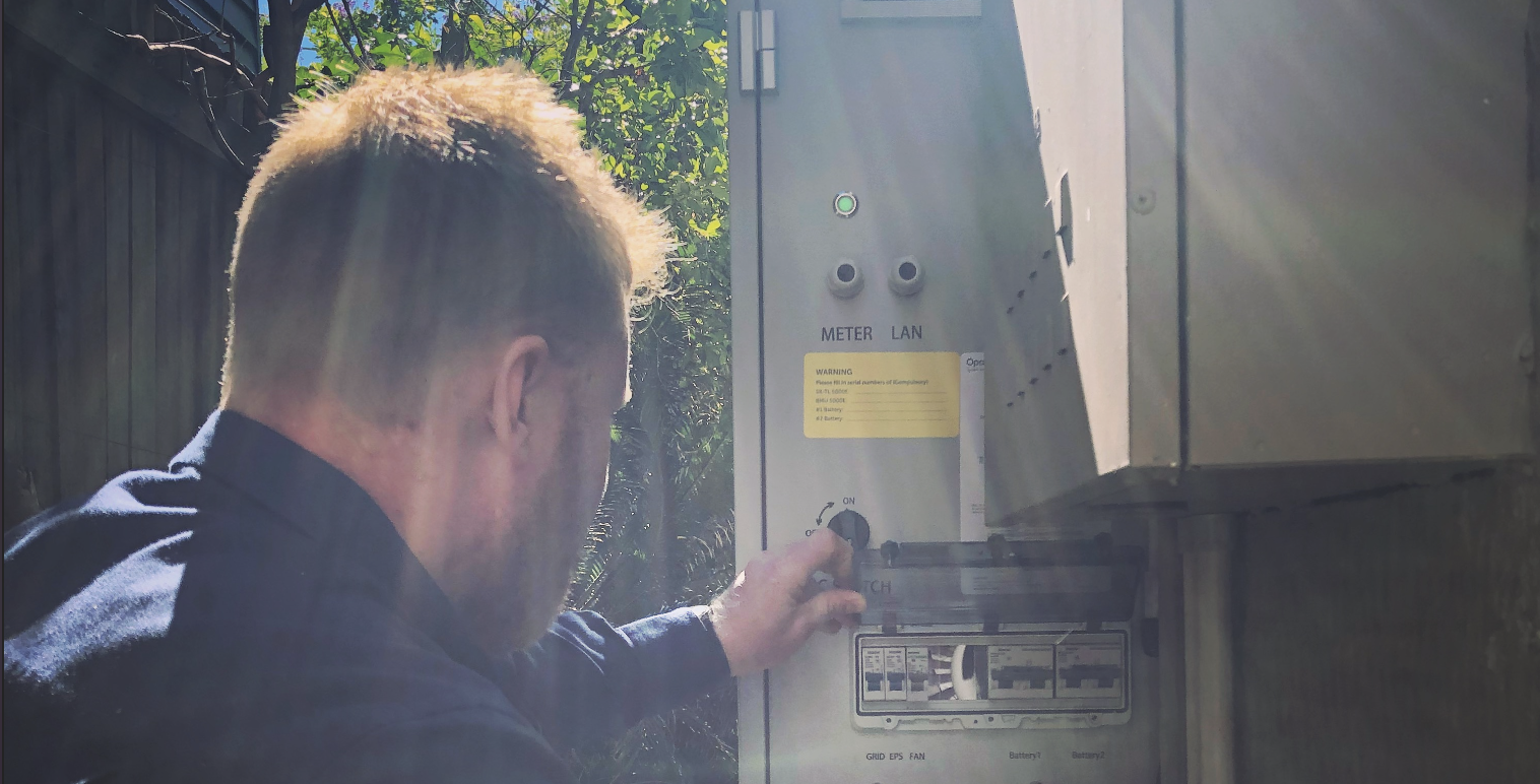 A man with blond hair wearing a dark shirt is adjusting a breaker switch box outdoors, with trees and sunlight visible in the background.