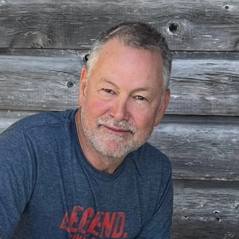 A middle-aged man with gray hair and a beard, wearing a blue t-shirt, sitting outdoors against a wooden background.
