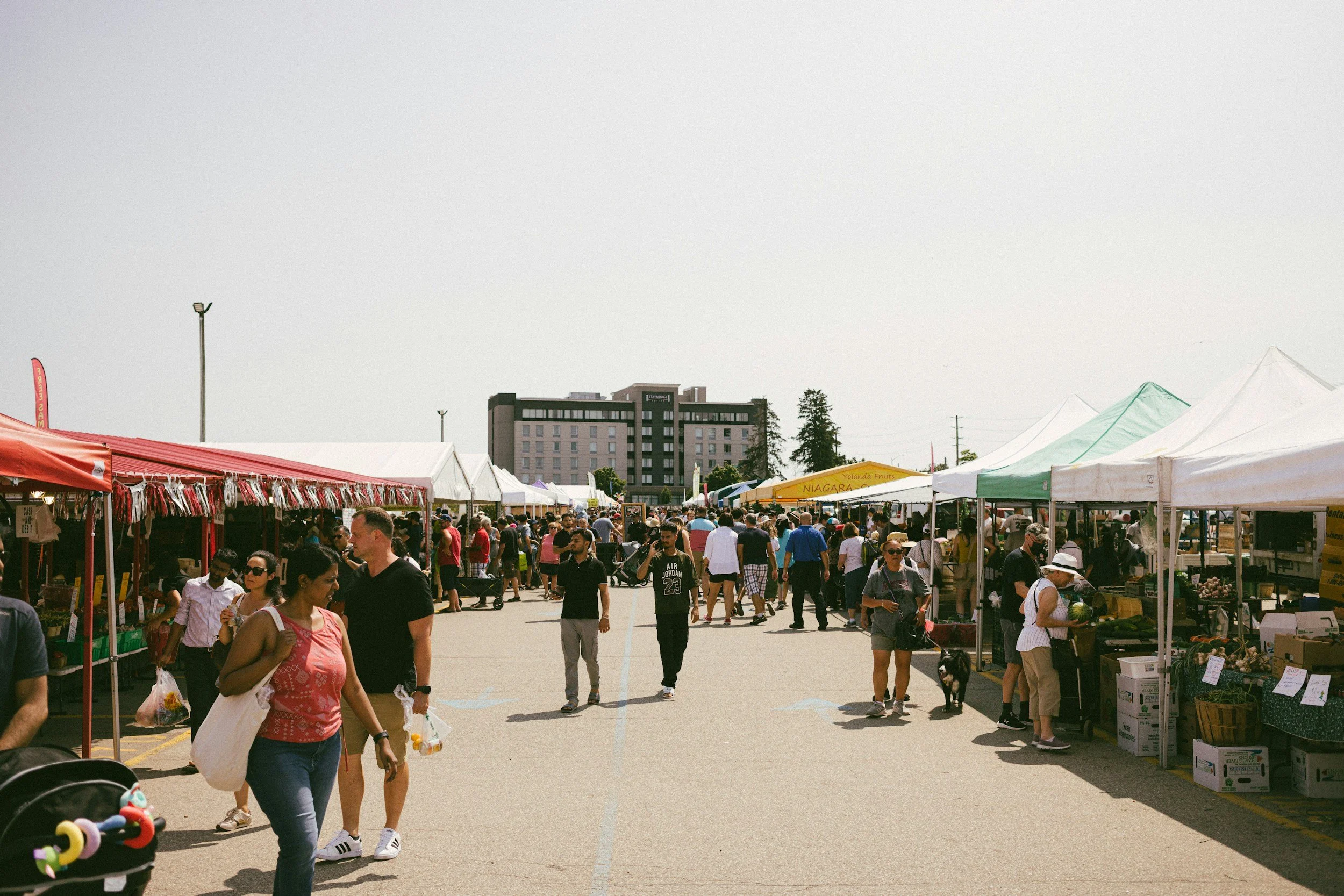 A crowded outdoor market with numerous tents and stalls selling goods. Many people are walking and browsing the market, some carrying bags. There is a large building and trees in the background under a clear sky.