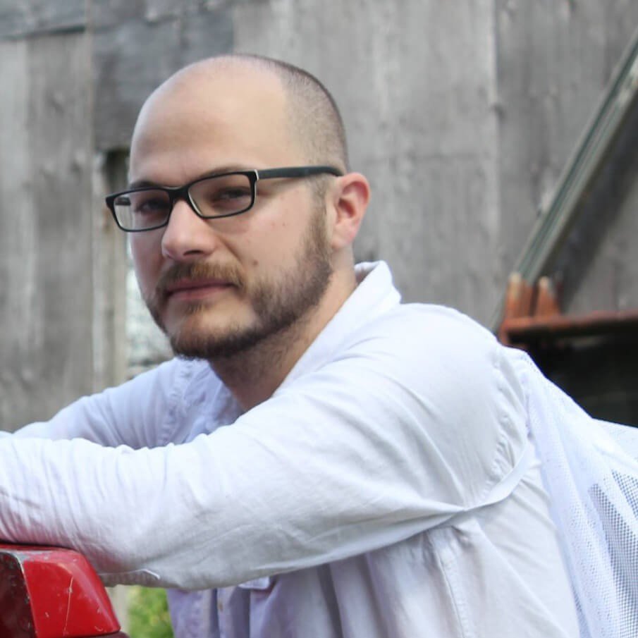 A man with glasses and a beard, wearing a white shirt, leaning on a red surface outdoors with a wooden fence background.