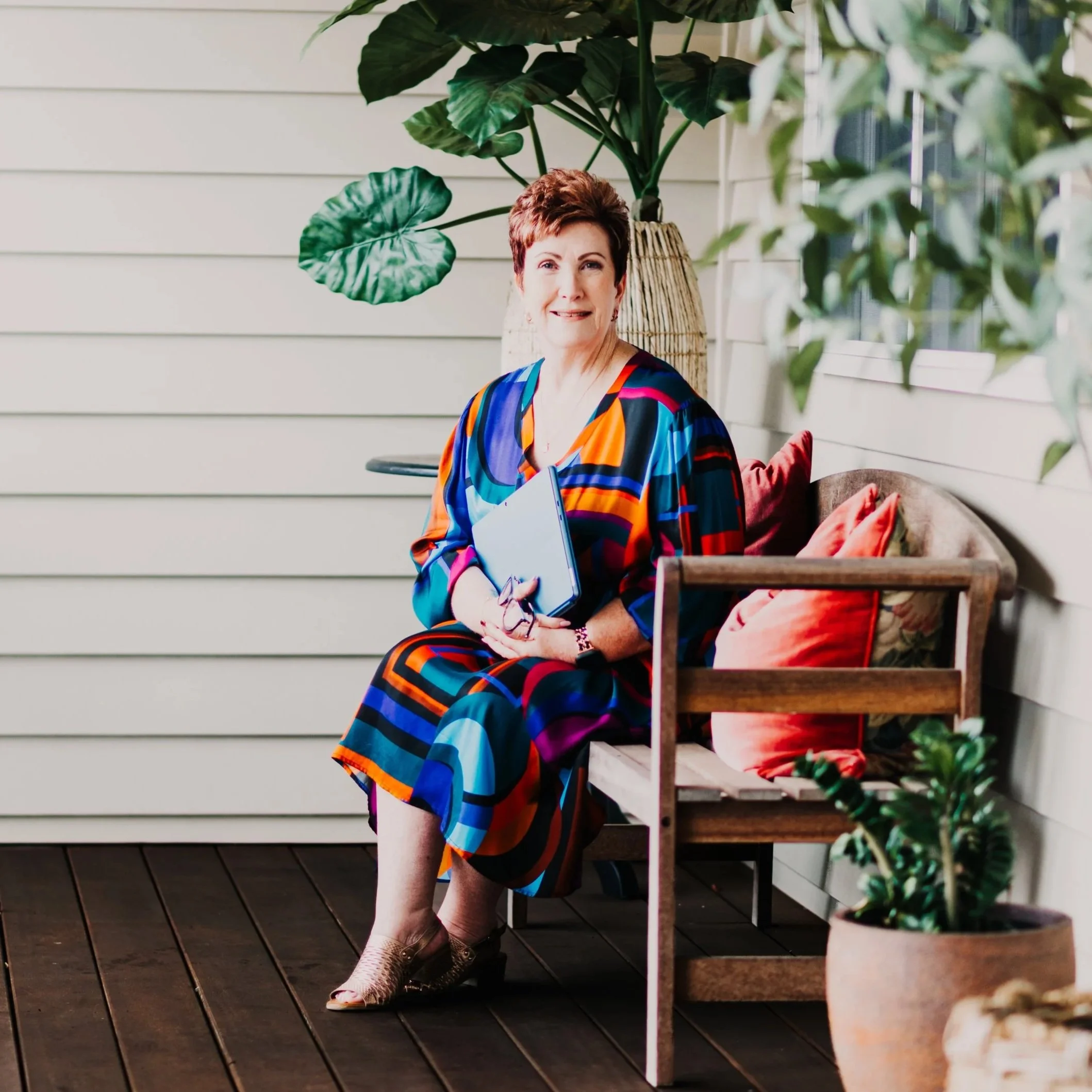 A woman with short reddish-brown hair wearing a colorful, striped dress sitting on a porch with white siding, next to a large potted plant with green leaves, holding a clipboard.