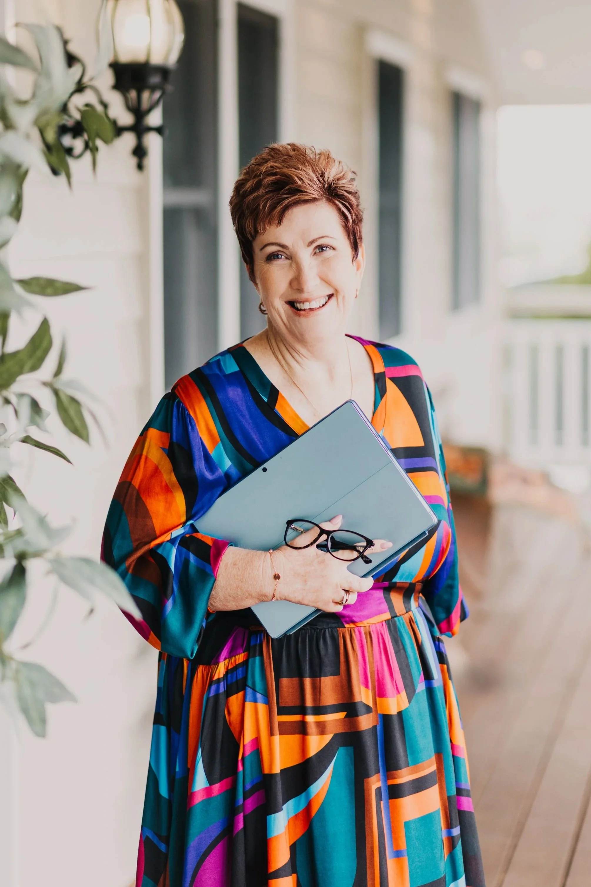 A smiling woman with short brown hair wearing a colorful, patterned dress, holding a tablet and glasses, standing on a porch with a white wall and windows in the background.