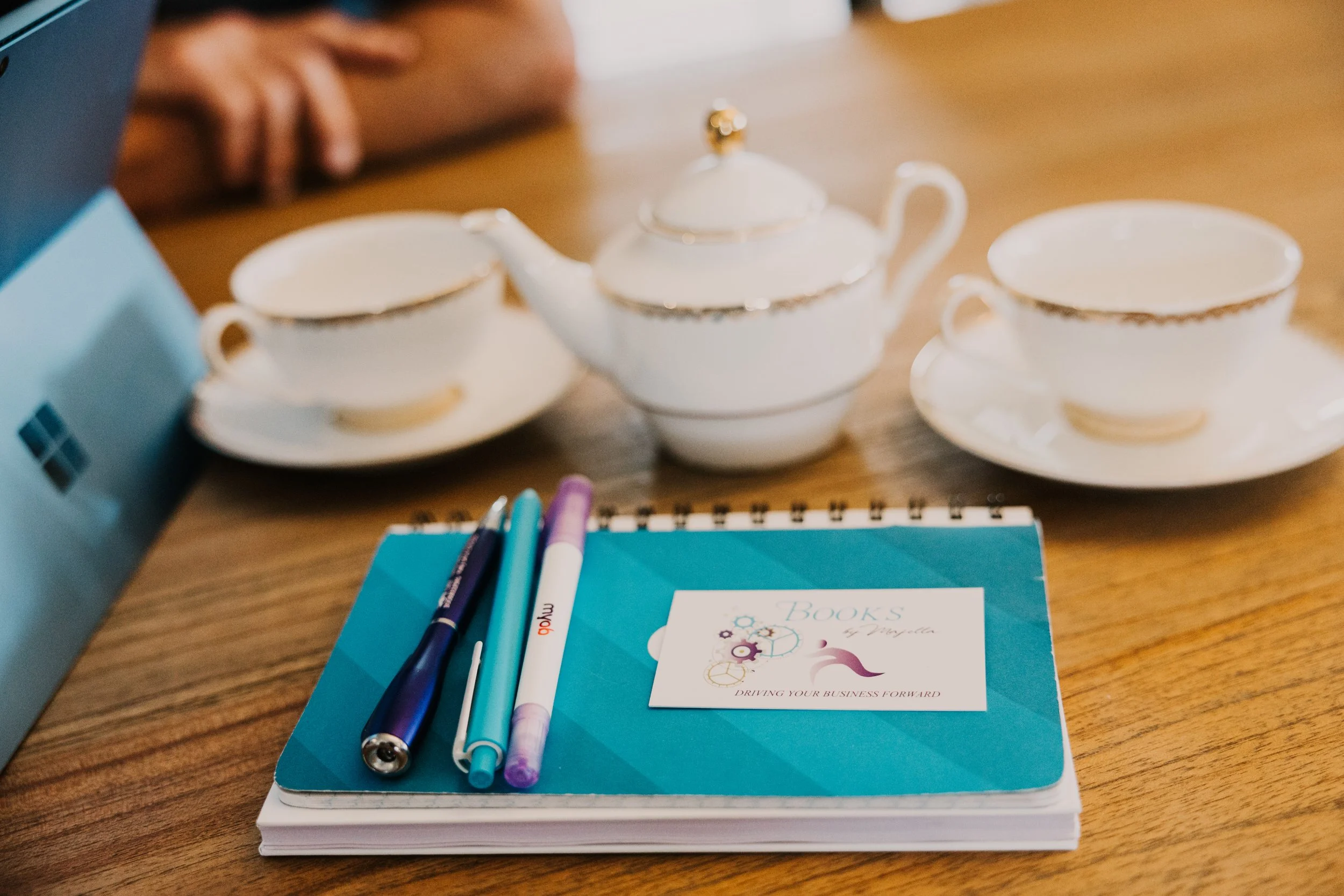 A wooden table with a white teapot and three matching teacups with gold trim. There are three pens and a blue notebook with a business brand card on top of it.