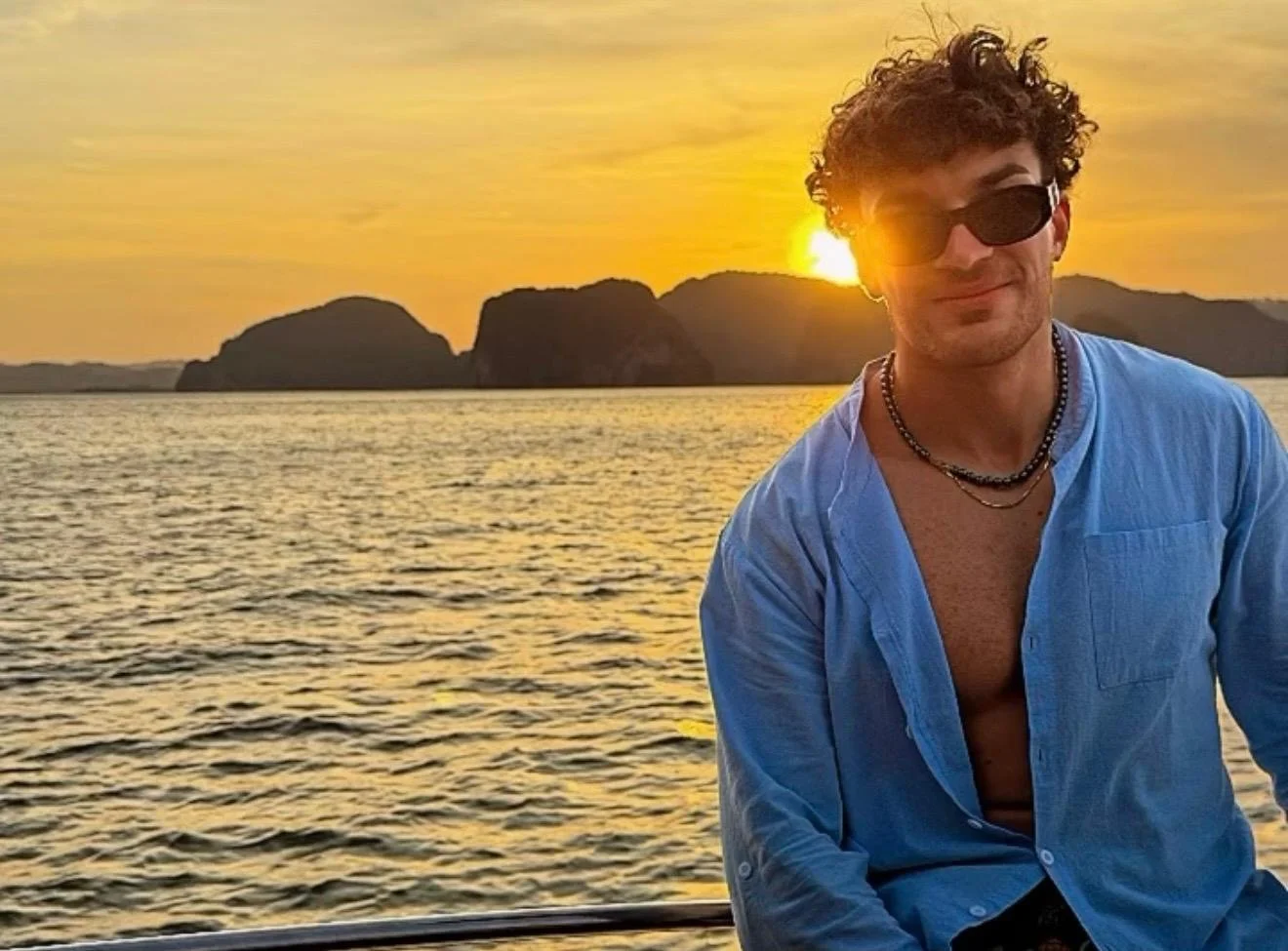 A man with curly hair wearing sunglasses, a blue unbuttoned shirt, and necklaces, smiling at sunset on a body of water with rocky islands in the background.