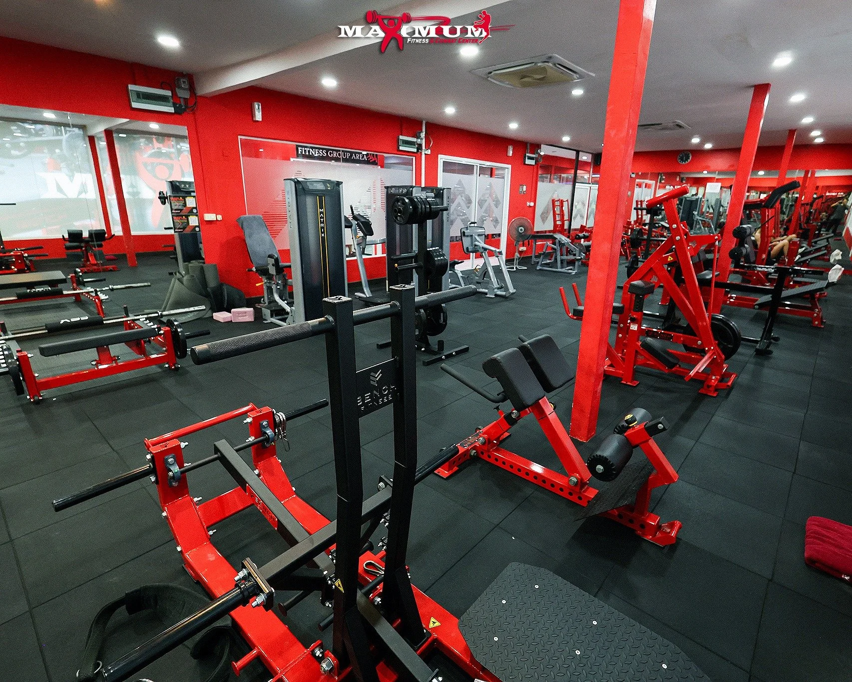 Interior of a gym with various red and black workout equipment, including weightlifting machines, benches, and cardio machines, with red walls and a black rubber floor.