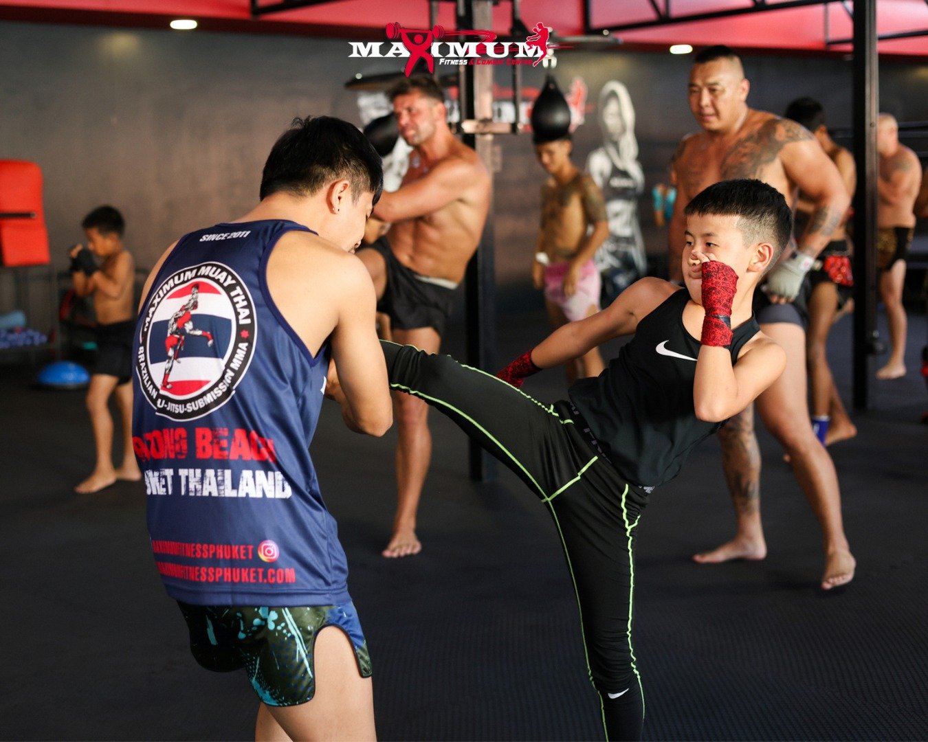 Two young boys practicing martial arts in a gym. One is performing a high kick while the other is blocking it. Several adults and children are in the background, some training and others observing. The gym has black flooring and red accents.