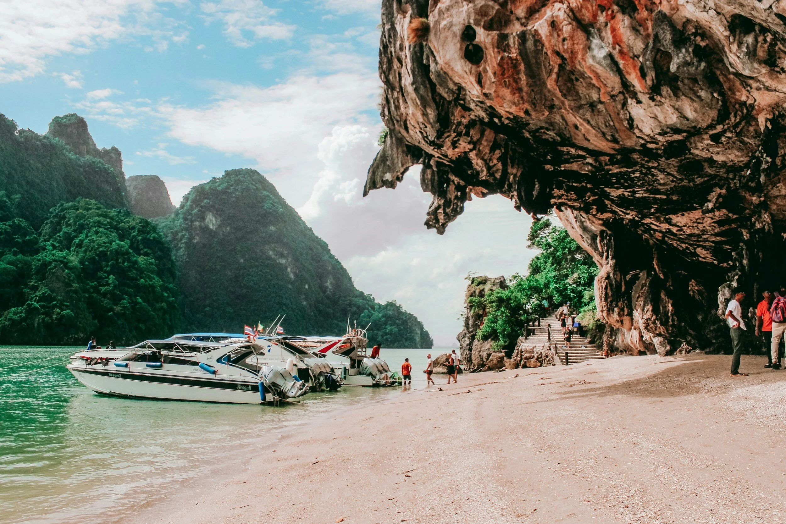 Tour boats docked on a sandy beach beside lush green cliffs under a partly cloudy sky, with people walking and exploring near the shoreline.