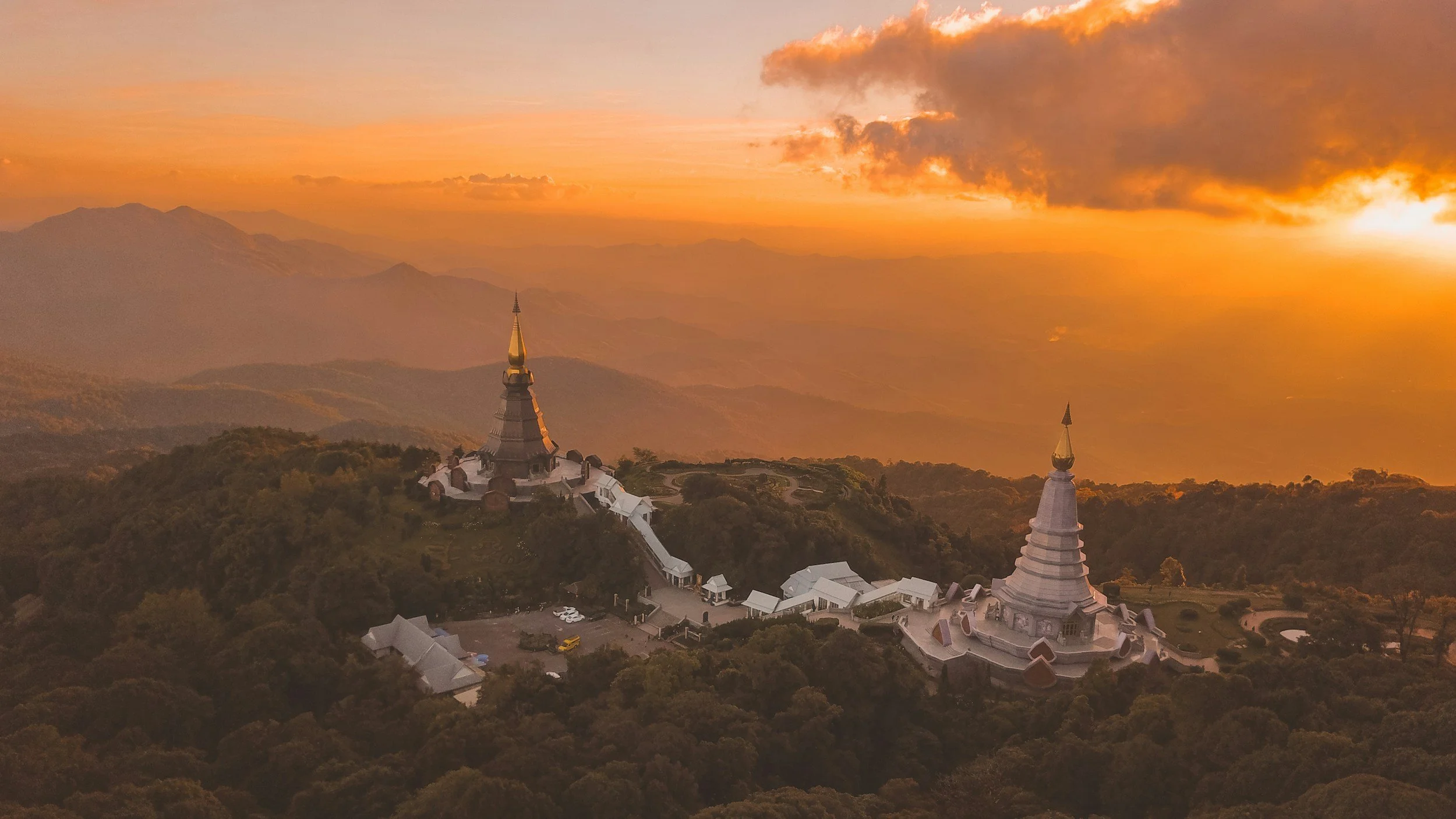 Two white and gold Buddhist stupas on a hilltop overlooking a mountainous landscape at sunset with orange and yellow sky and clouds.