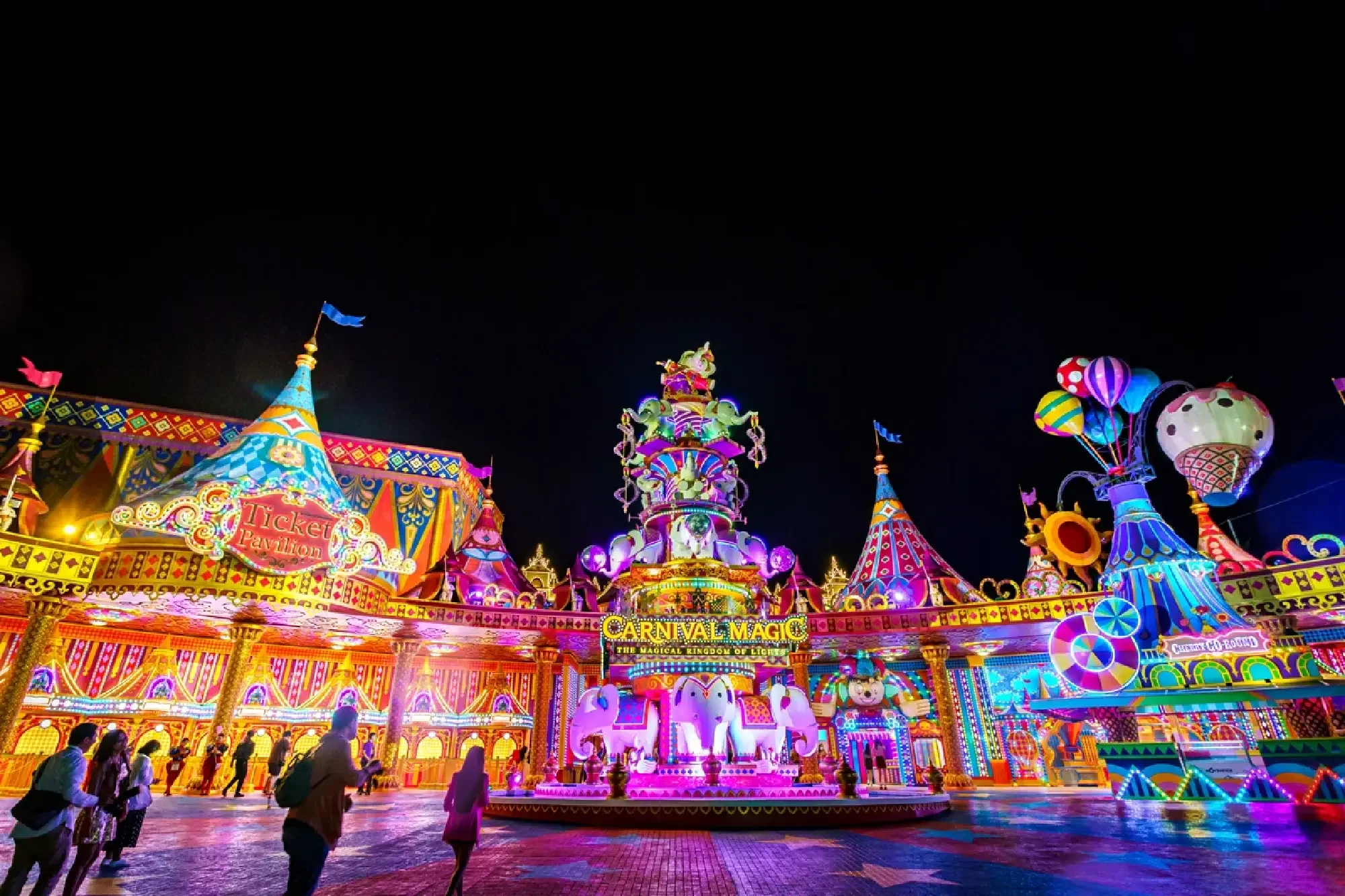 Colorful carnival amusement park at night with bright lights, rides, decorations, and people walking around.