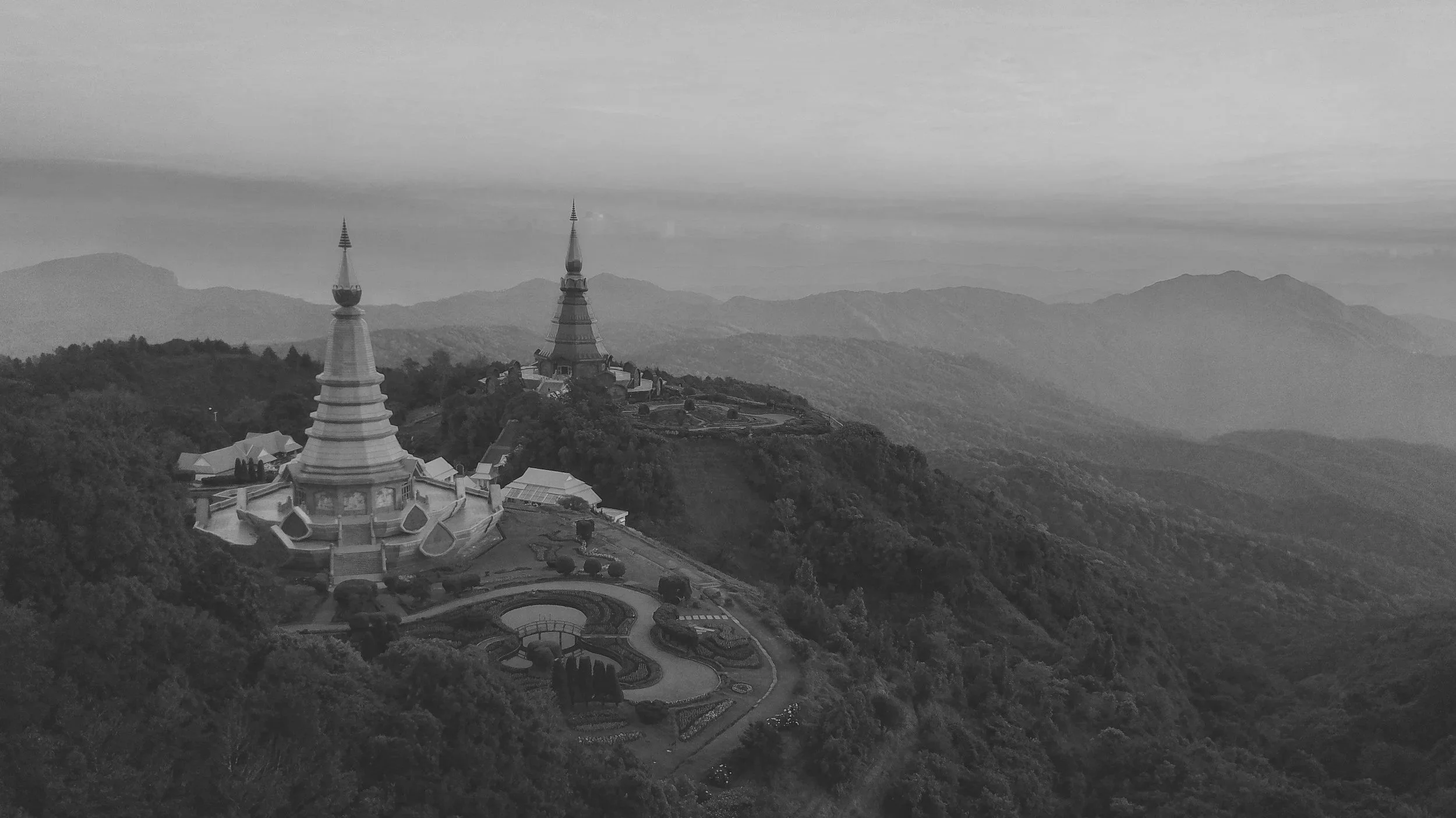 Black and white photo of a temple complex on a hilltop with multiple spires, surrounded by lush trees and rolling mountains in the background.