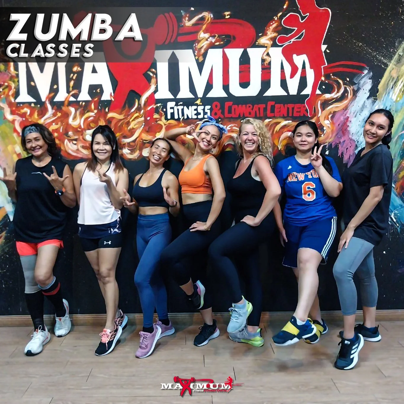 Group of seven women posing in front of a colorful wall with zumba class advertisement, wearing athletic clothing and smiling.