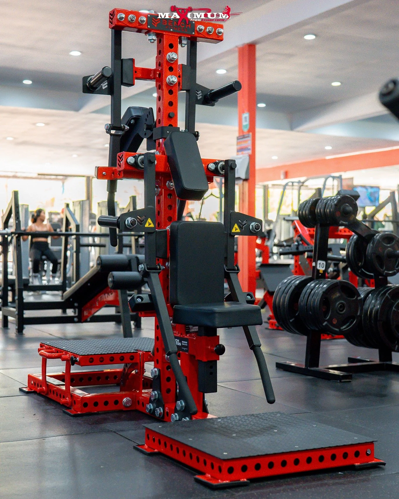 Red and black strength training machine with padded seat and various attachments, in a gym with weight plates and other exercise equipment in the background.